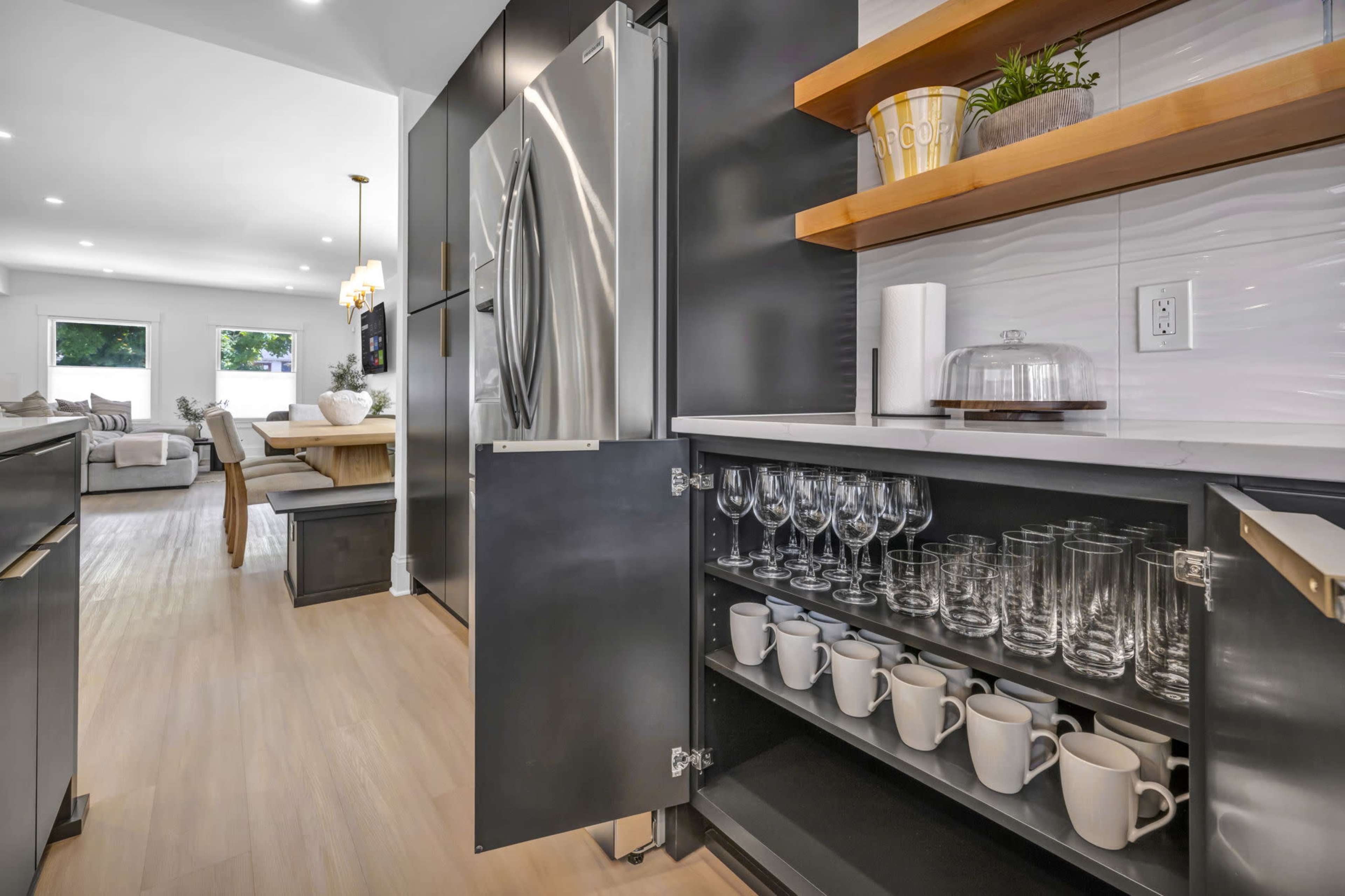 A modern kitchen features a stainless steel refrigerator, open shelving with decorative items, and a cabinet displaying glassware and mugs.