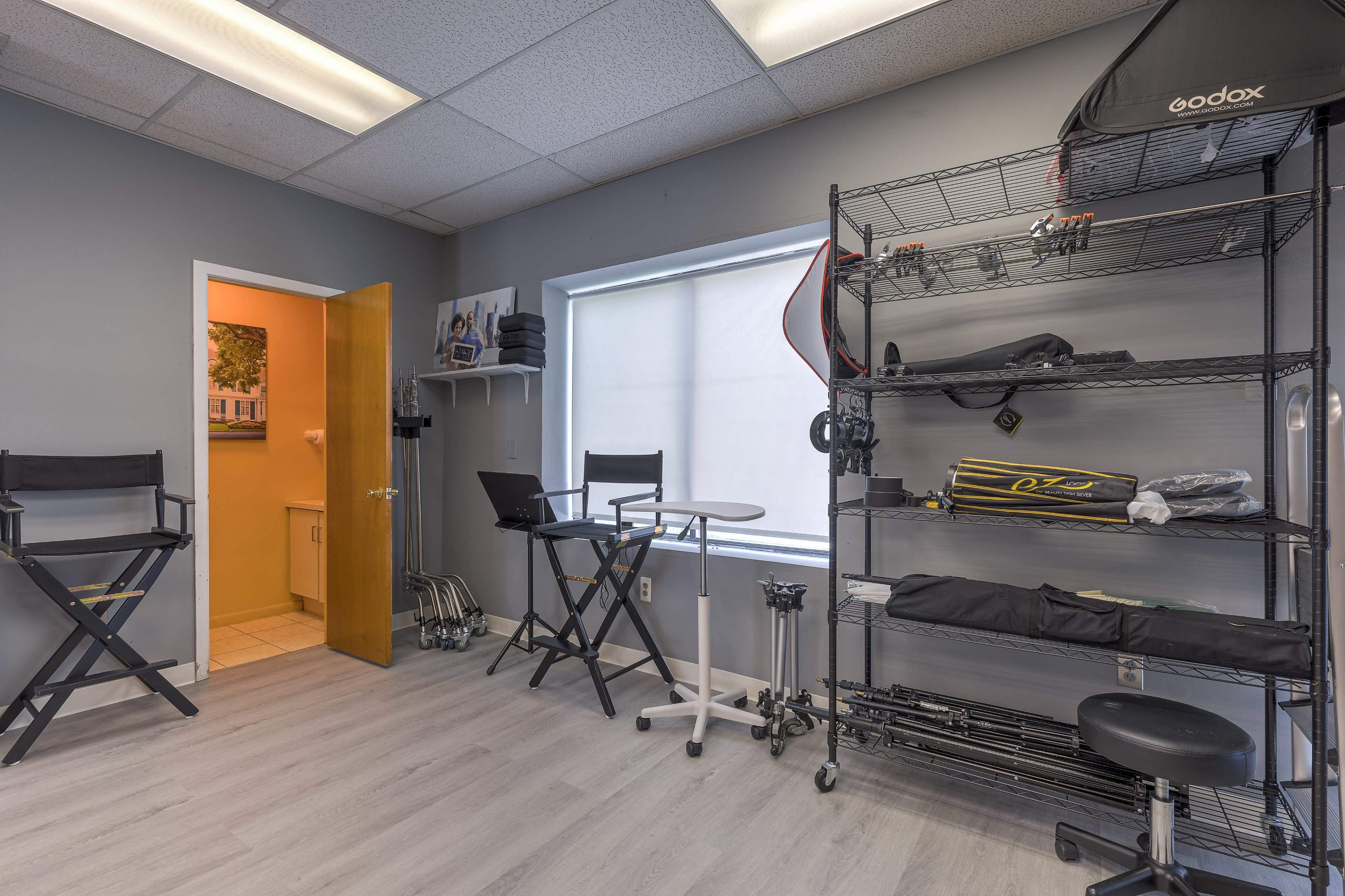A small photography studio with shelves holding equipment, two director's chairs, and a doorway leading to another room.