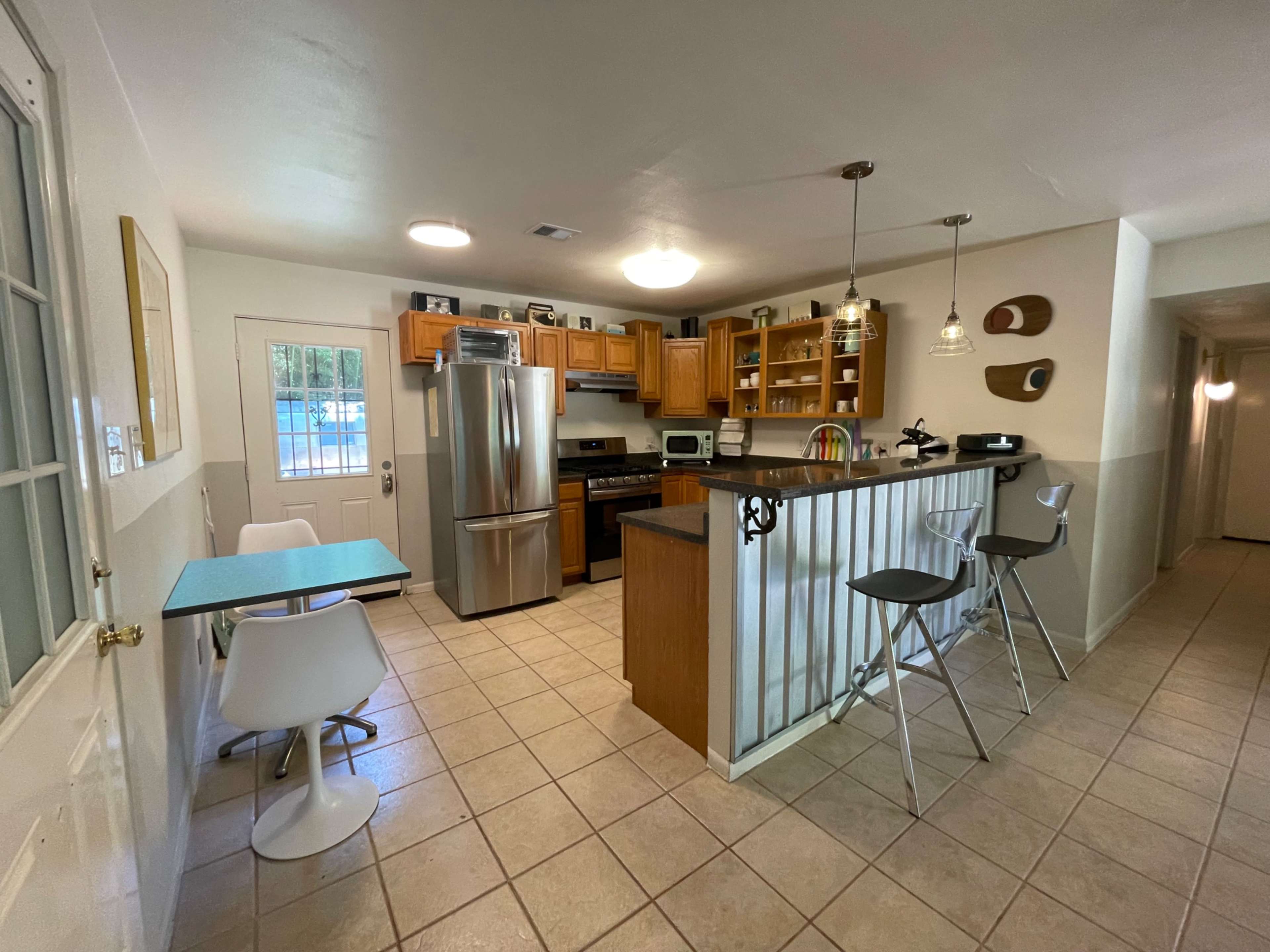 The image shows a compact kitchen with wooden cabinets, a stainless steel fridge, and a small dining area featuring a table and chairs.