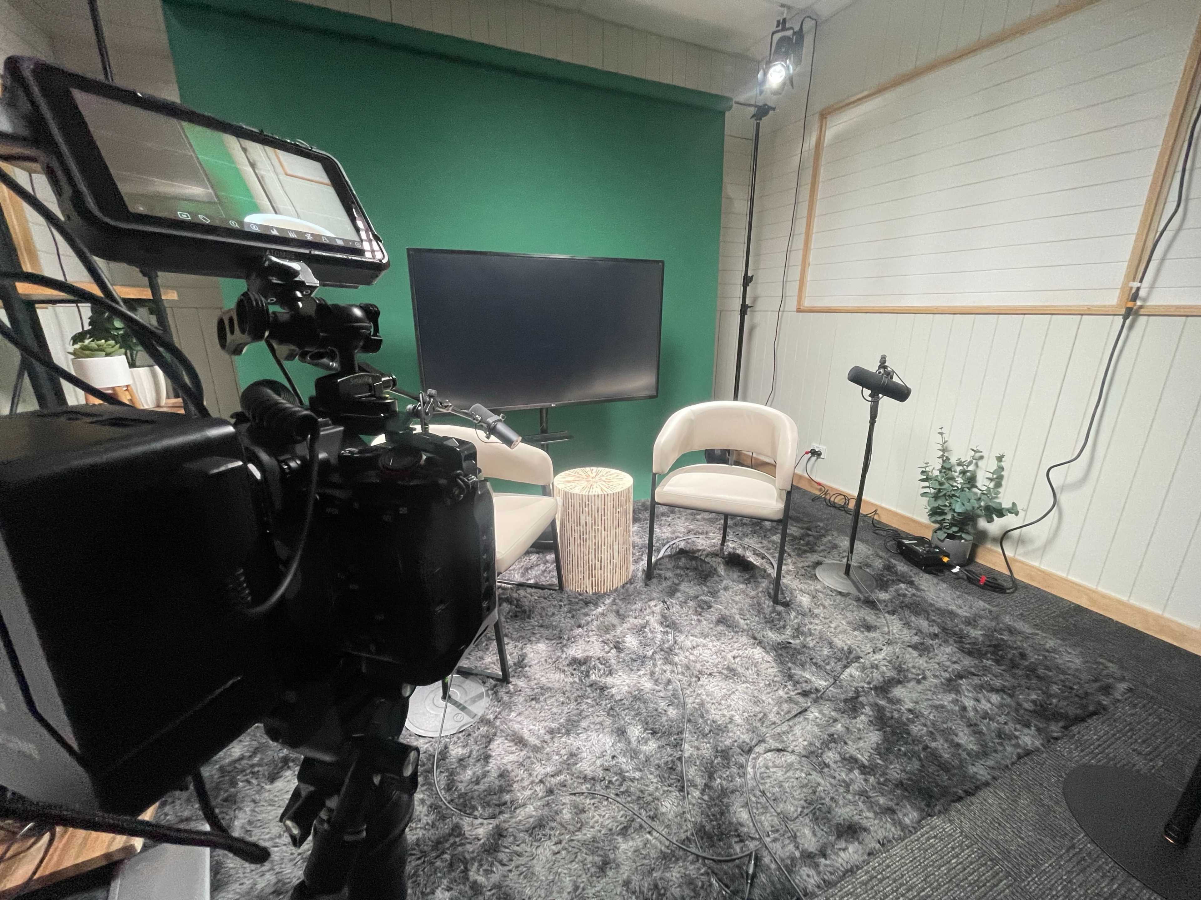 The image shows a small studio setup with two chairs, a table, and a television screen against a green backdrop, along with various lighting and camera equipment.
