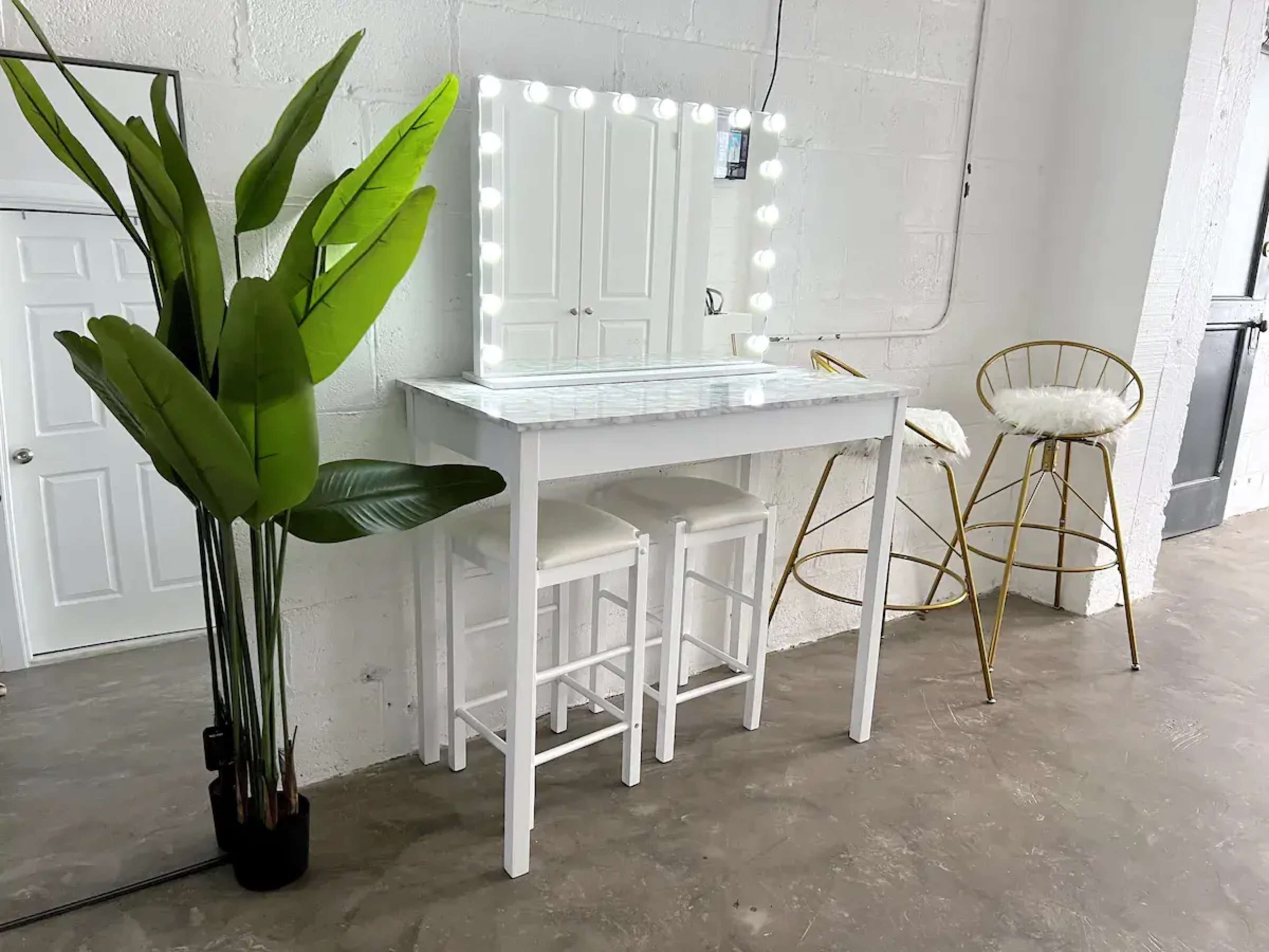 A white vanity table with a mirror surrounded by lights, two white stools, and a tall gold bar stool, set against a plain wall with a large plant in the foreground.