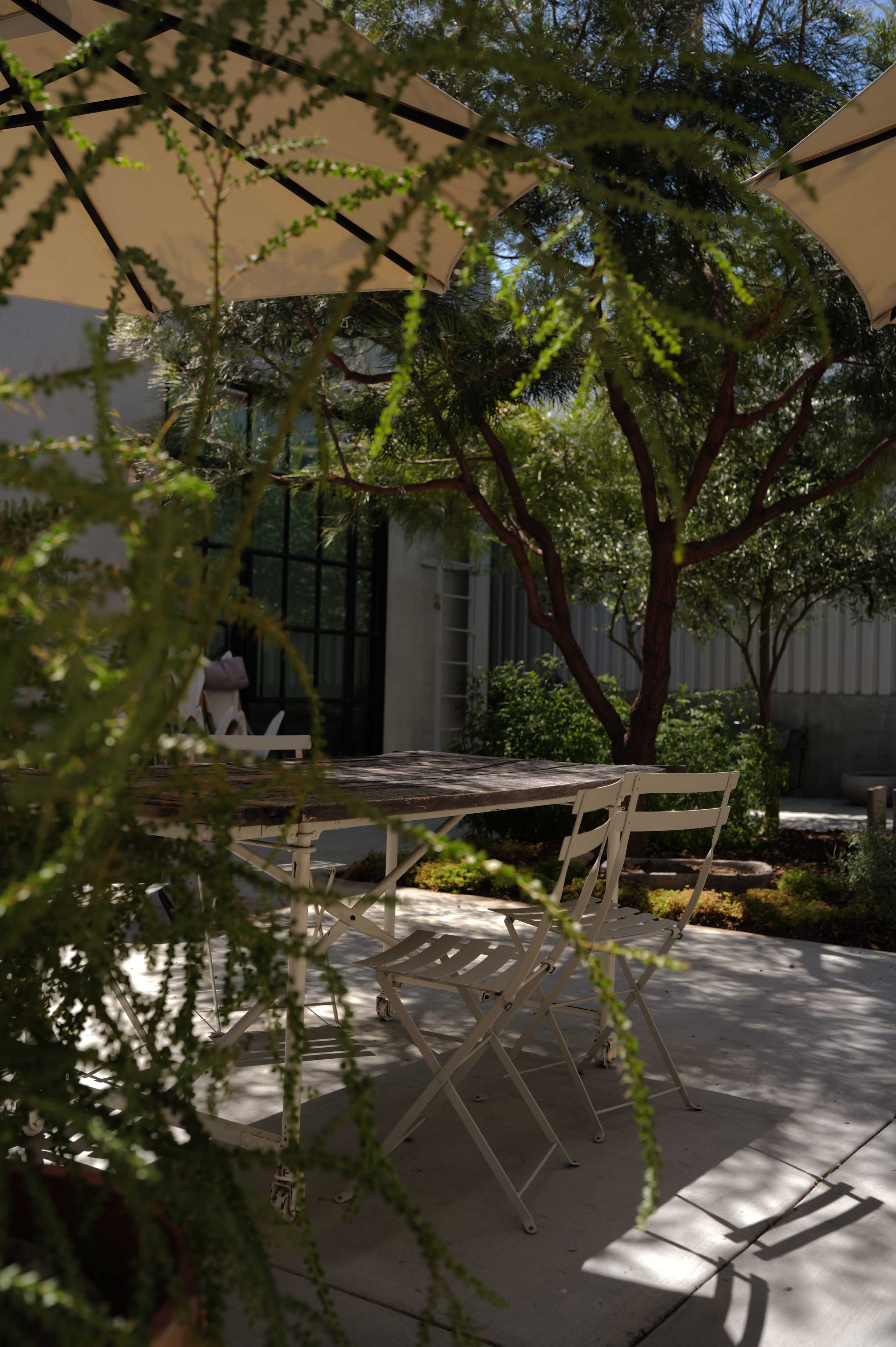 A small outdoor dining area features white chairs placed around a wooden table, surrounded by various plants and shaded by umbrellas.