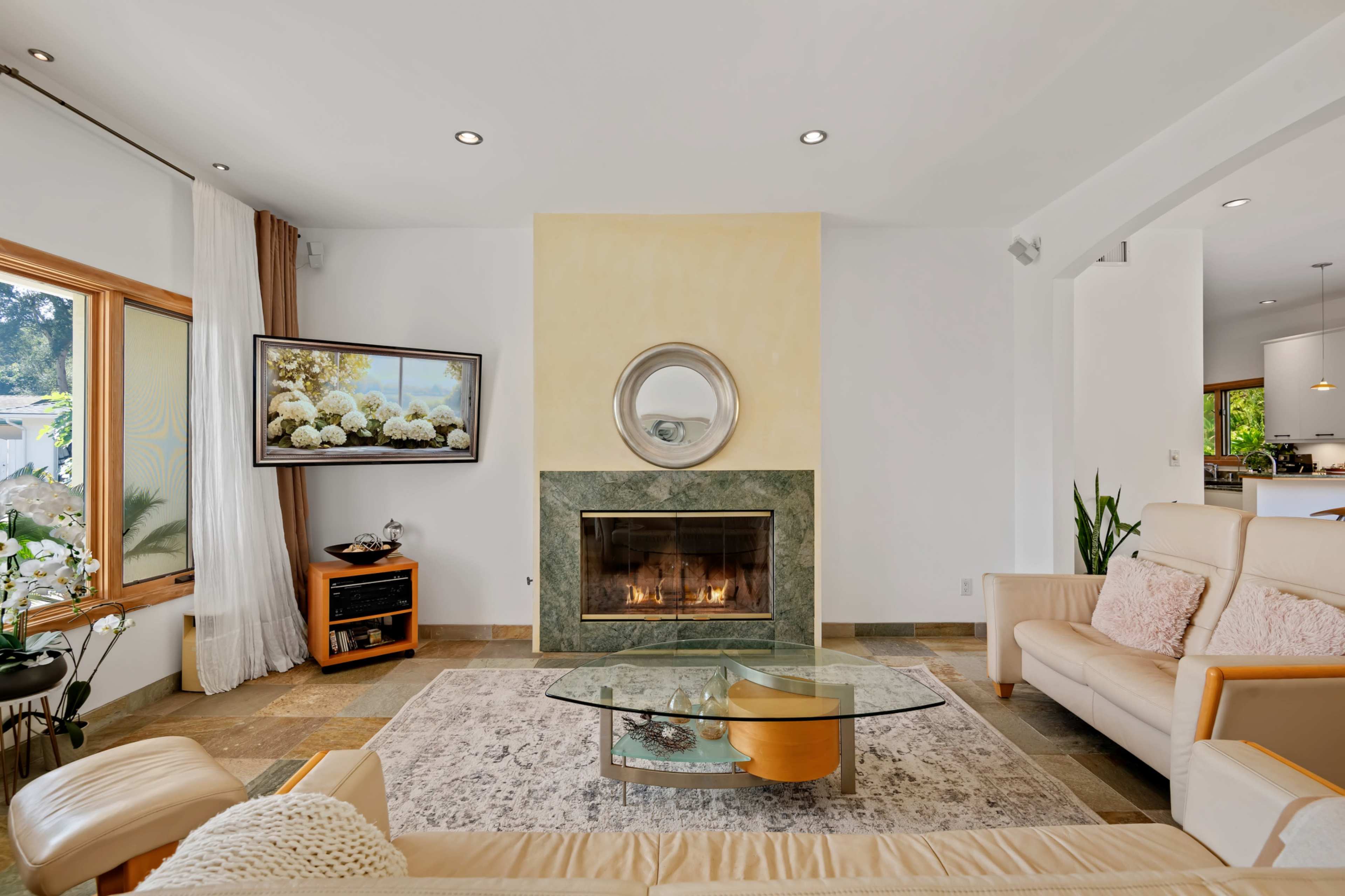 The image shows a modern living room featuring a central fireplace with a mirror above it, a television mounted on the wall, and light-colored furniture arranged around a glass coffee table on a patterned rug.