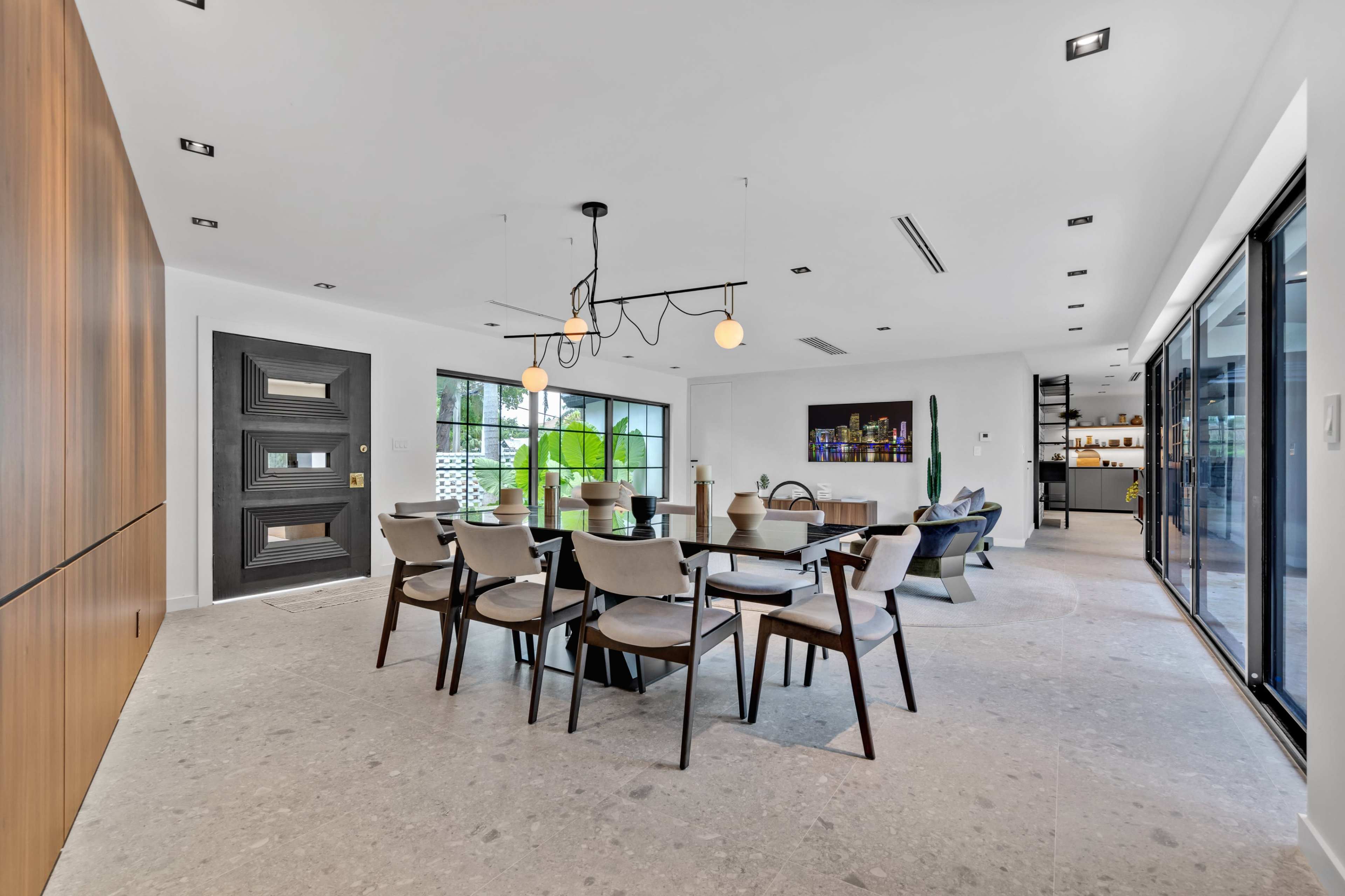 A modern dining area with a large table surrounded by chairs, a stylish light fixture overhead, and large windows providing natural light.