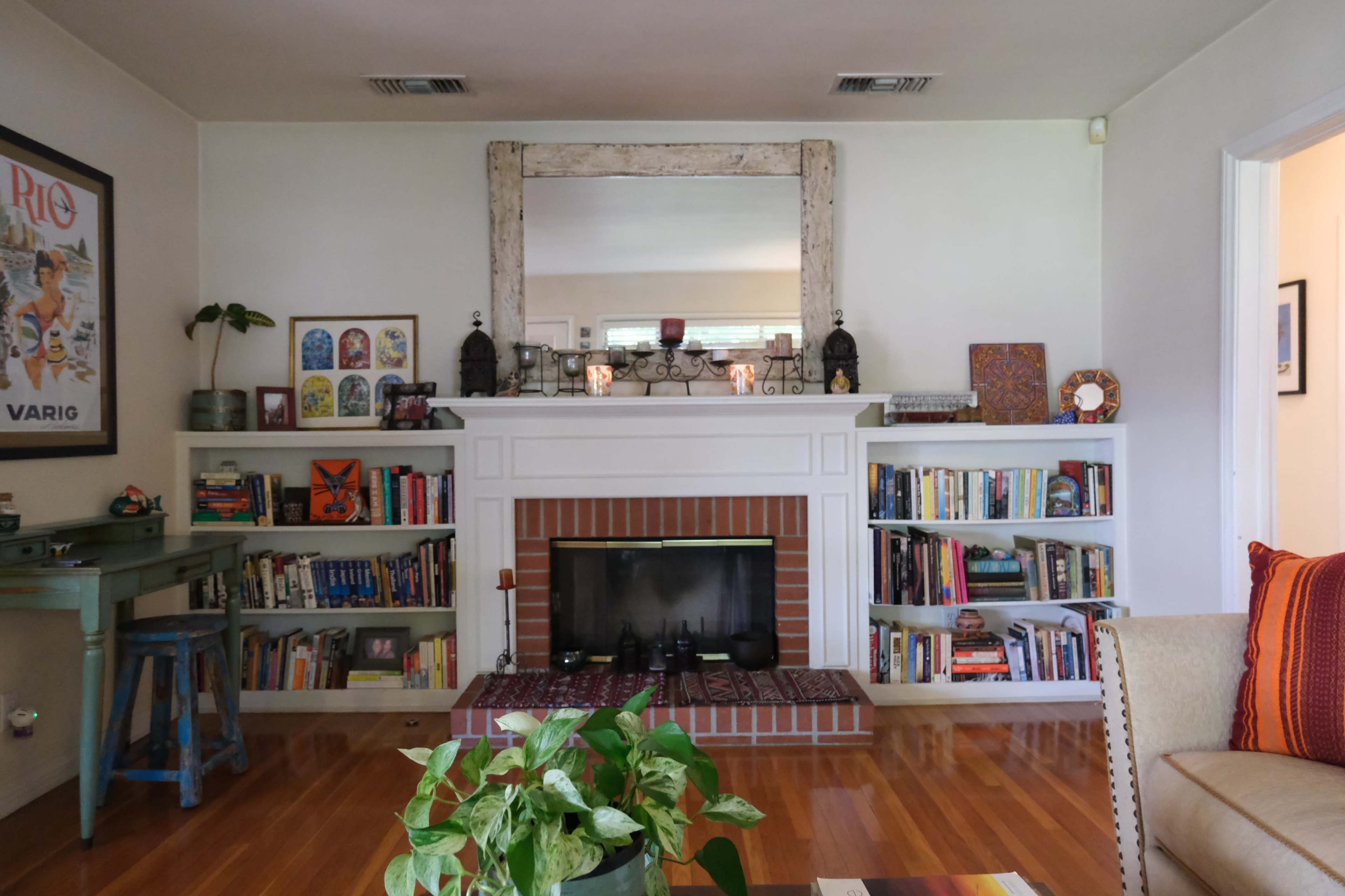 The image shows a cozy living room featuring a fireplace with a large mirror above it, flanked by bookshelves filled with books and decorative items, and a seating area with colorful cushions.