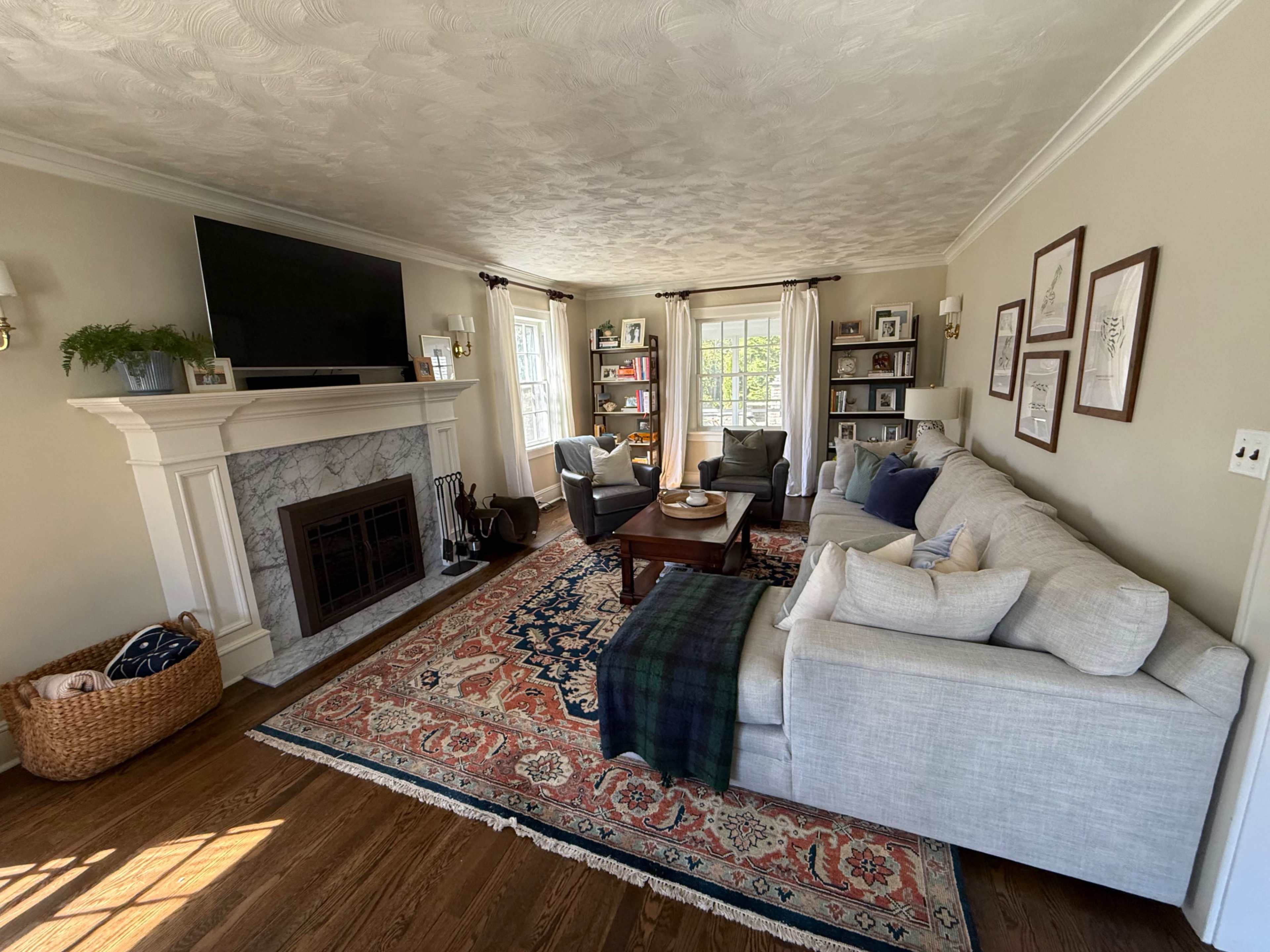 The image shows a cozy living room with a light-colored sectional sofa, a coffee table, a large television above a marble fireplace, bookshelves, and natural light coming through the windows.