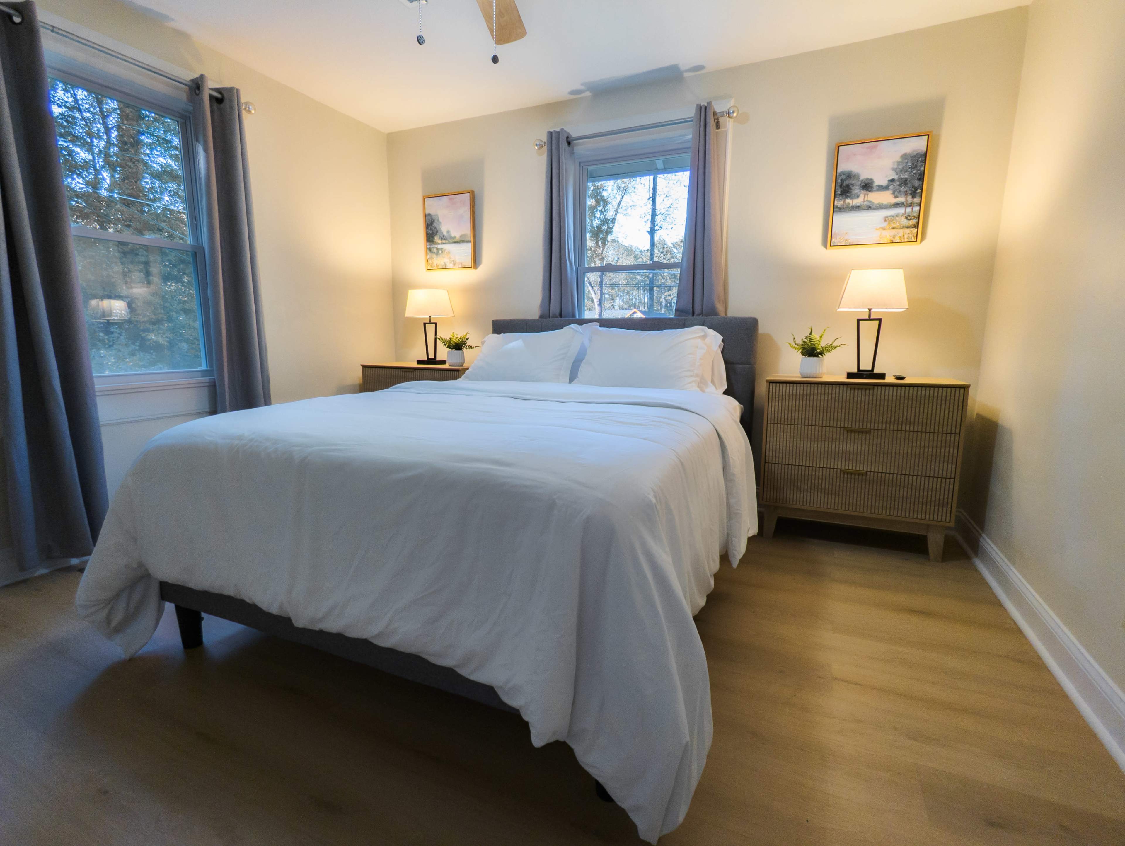 A neatly made bed with white linens is centered in a well-lit bedroom featuring nightstands with lamps on either side and framed artwork on the walls.