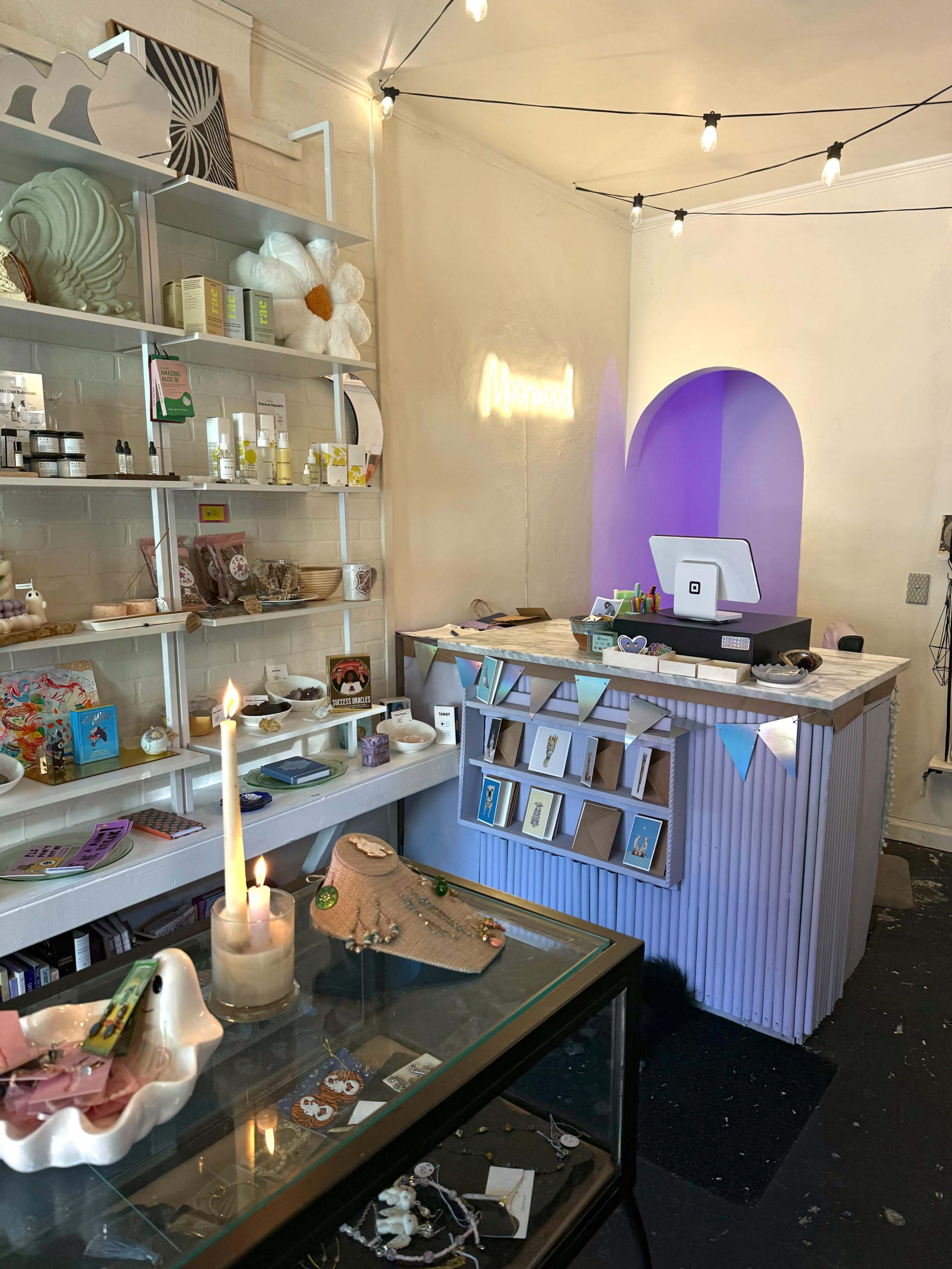 The image shows a retail space featuring a countertop with a point-of-sale system, shelves stocked with various products, and decorative elements like a candle and string lights.