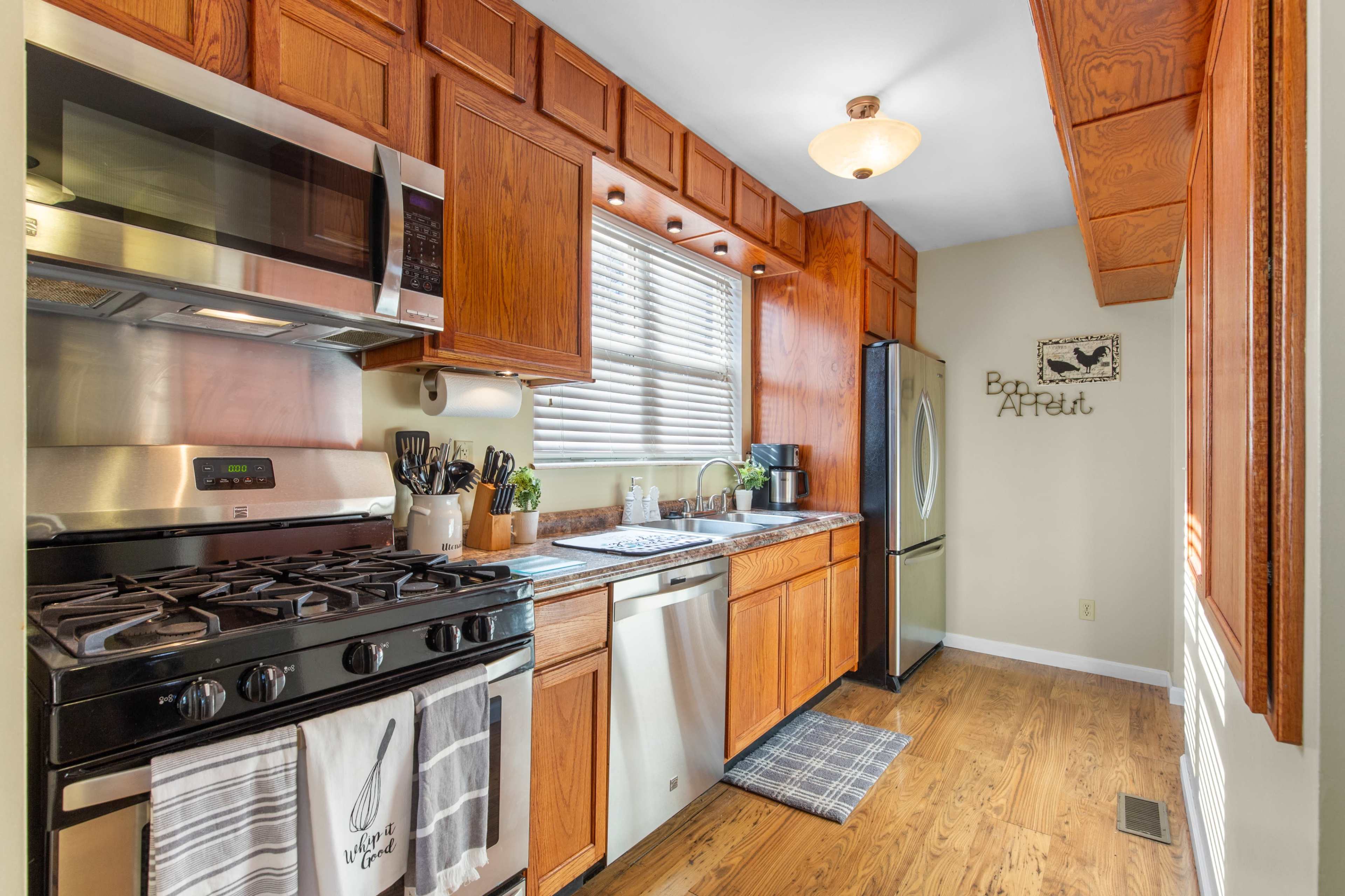 The image shows a kitchen with wooden cabinets, stainless steel appliances, and a window providing natural light.