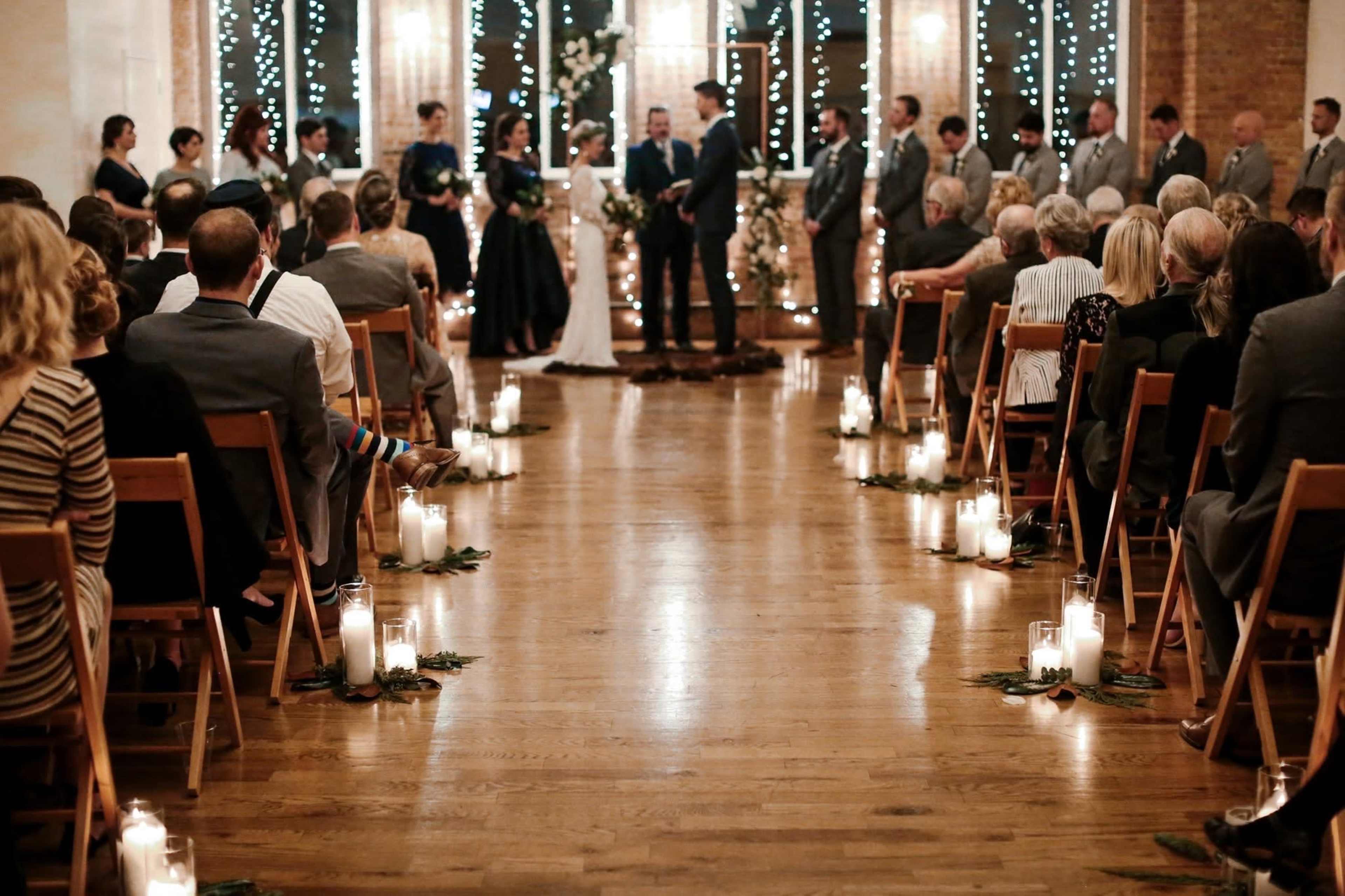 A wedding ceremony takes place indoors with guests seated in rows, surrounded by candlelight and decorated with greenery.