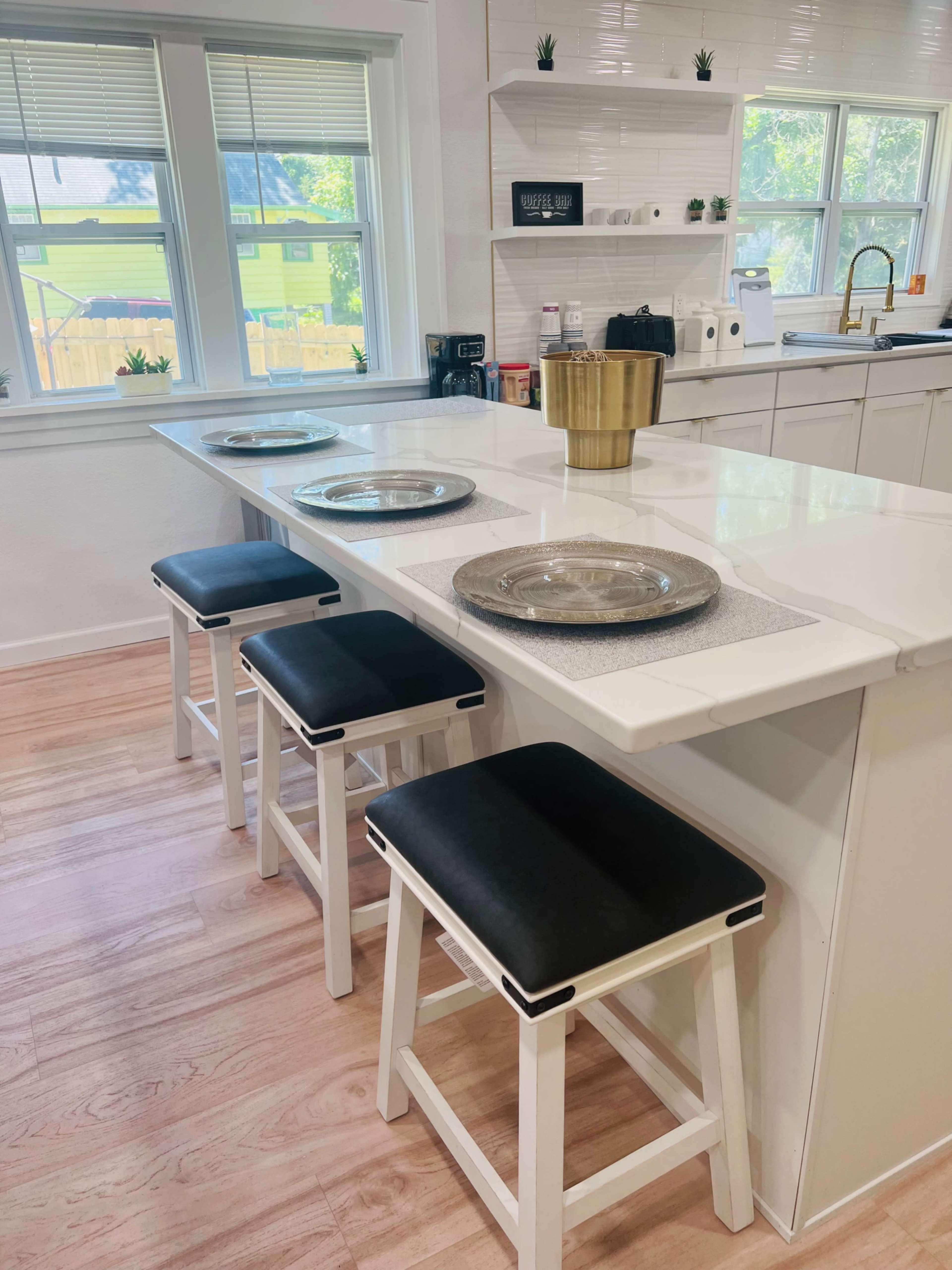 A modern kitchen features a white countertop island with four black and white stools, set with decorative plates and a gold bowl.