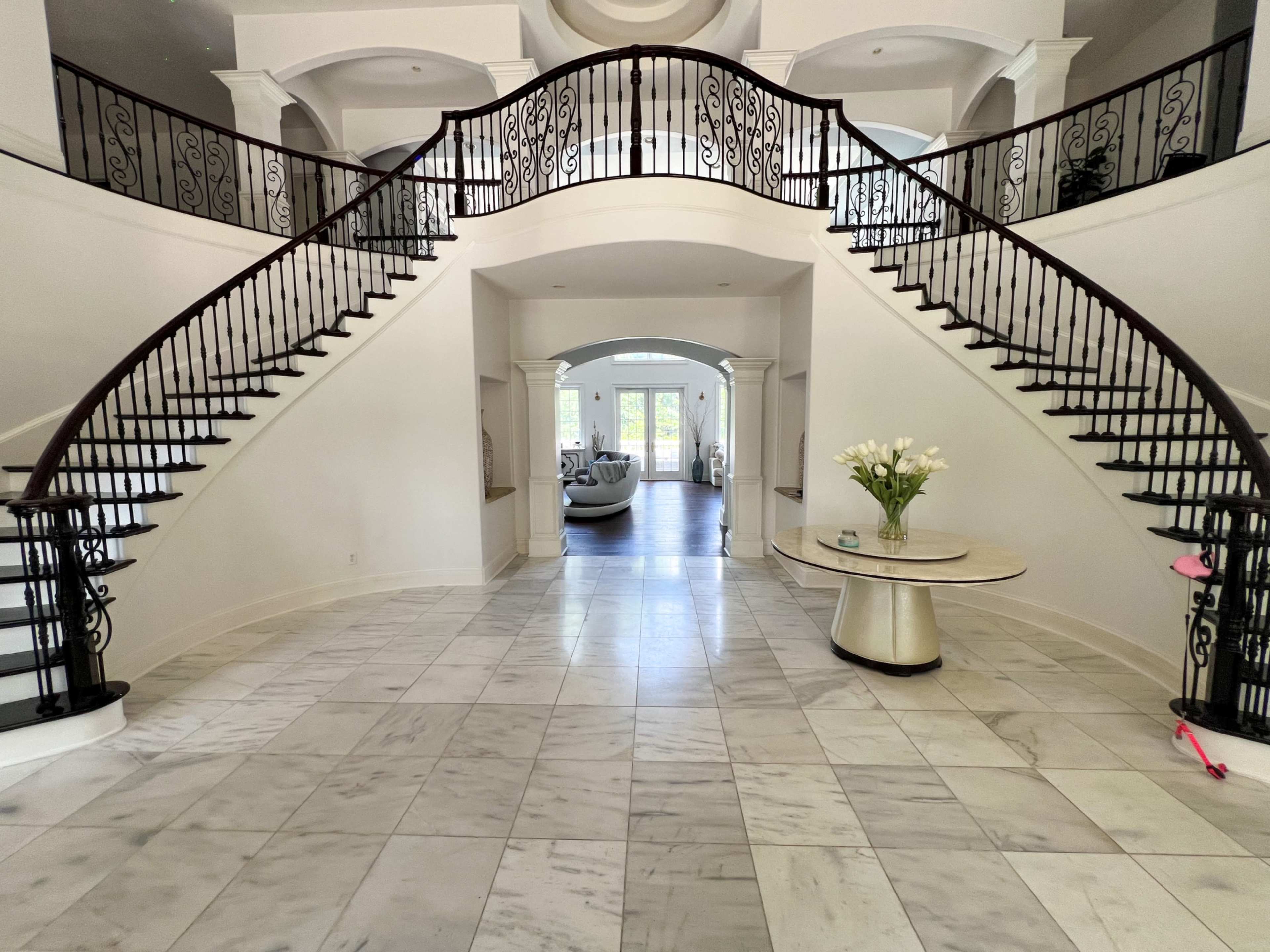 A grand foyer features a double staircase with black wrought iron railings, leading to an open living space beyond.