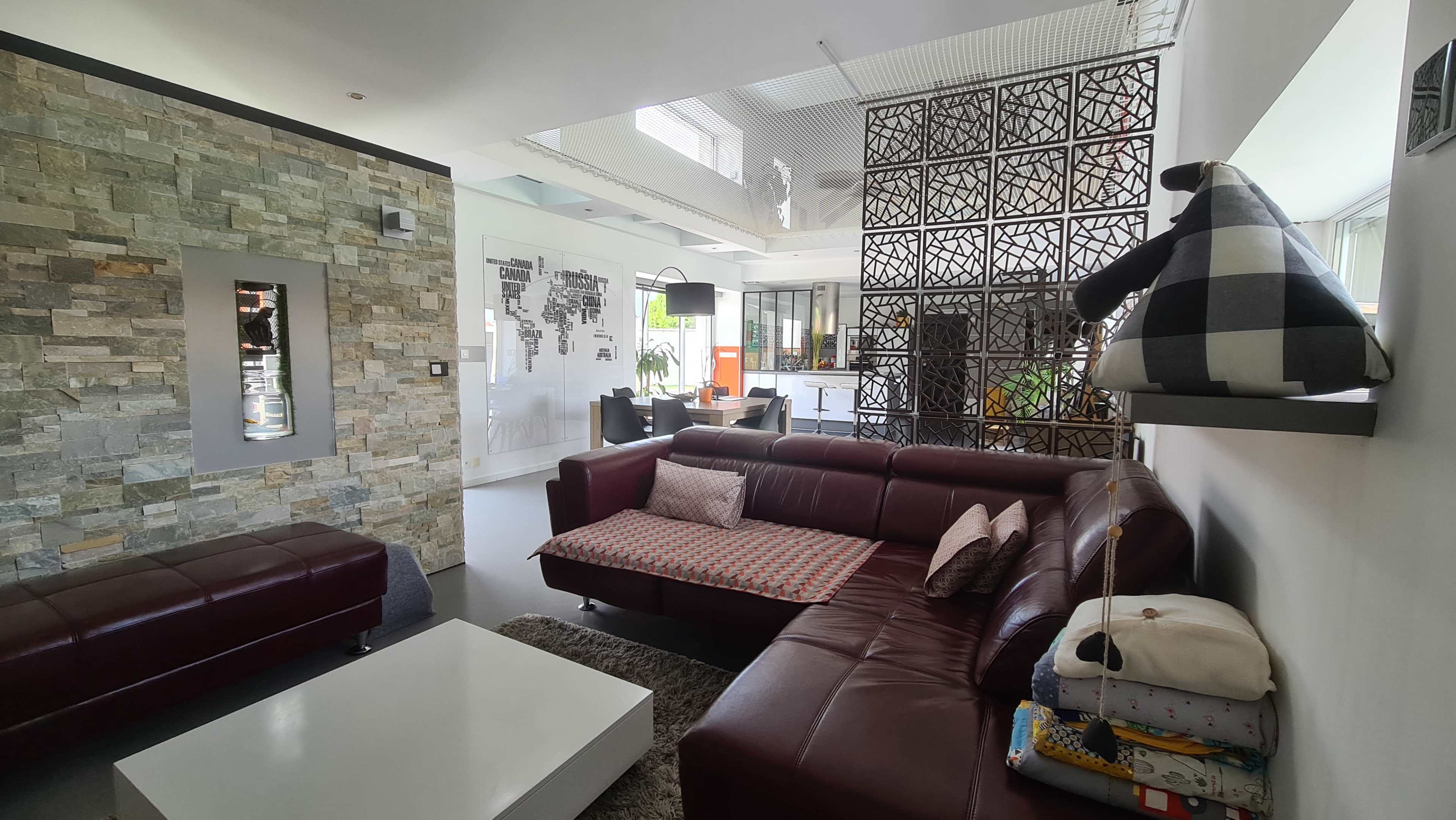 The image shows a modern living room with a maroon leather sofa, a stone-accent wall, and a glass partition leading to a dining area.