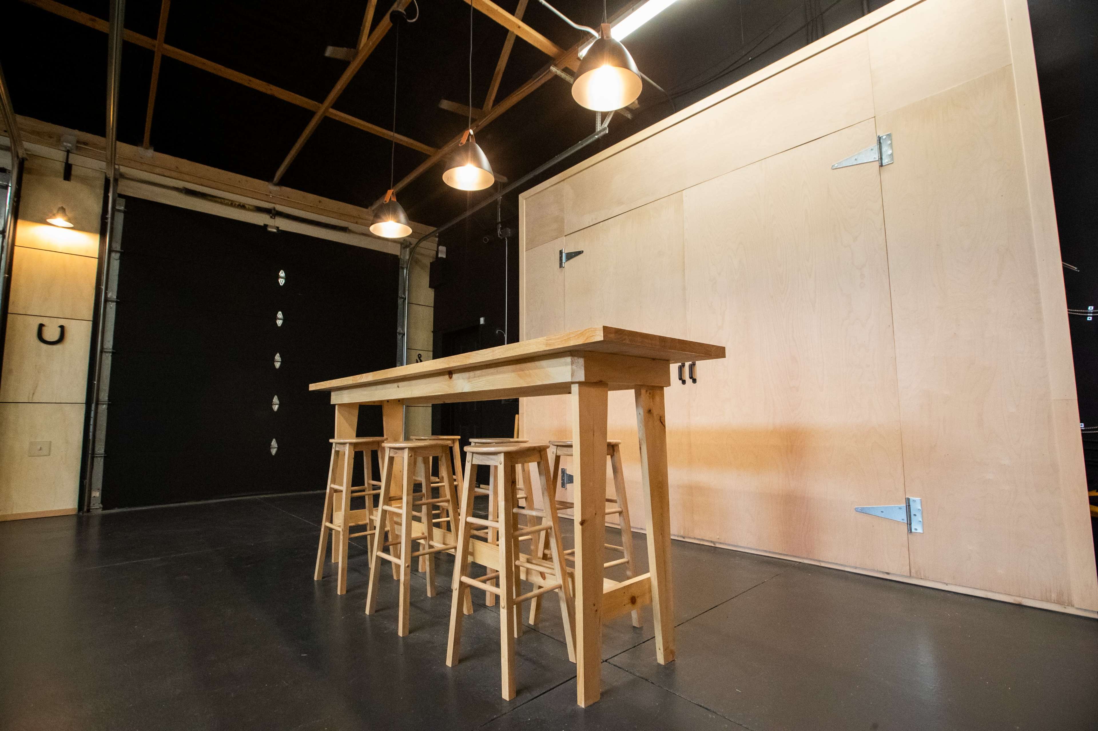 The image shows a minimalist studio space featuring a wooden table with four high stools in front of a large, light-colored panel and a dark garage-style door.