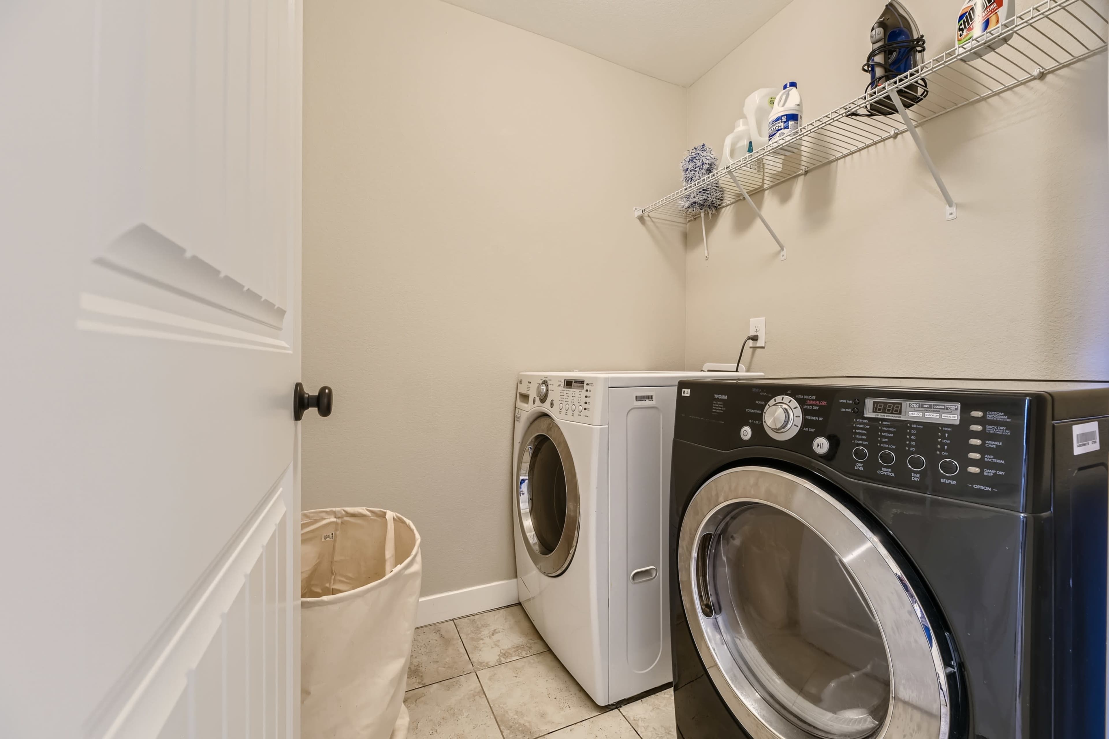 A laundry room containing a white washing machine and a black dryer, with a wire shelf above holding various laundry supplies and a beige laundry basket on the floor.