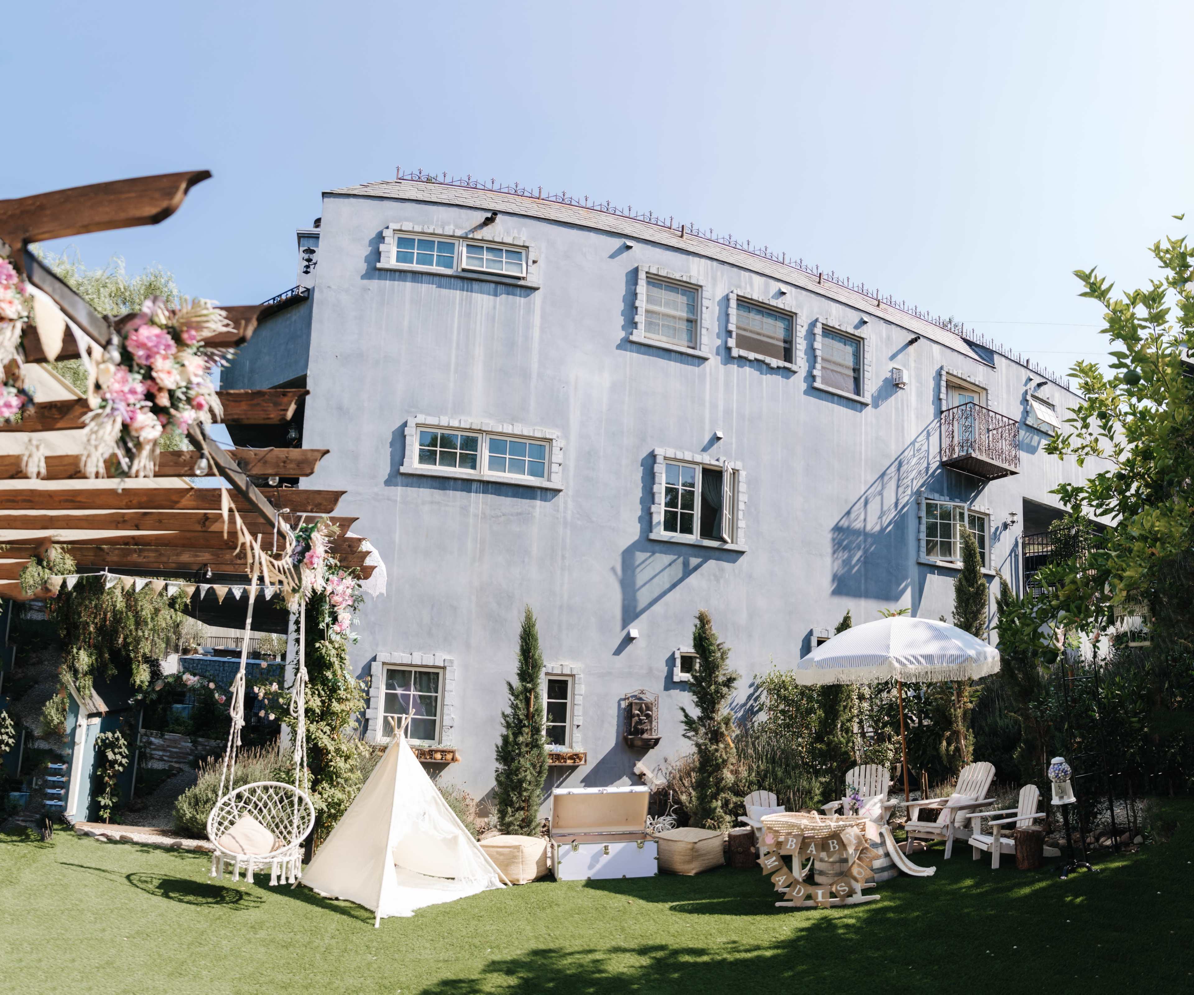 A blue building with multiple windows and balconies sits behind a landscaped yard featuring a swing, tent, and patio furniture.