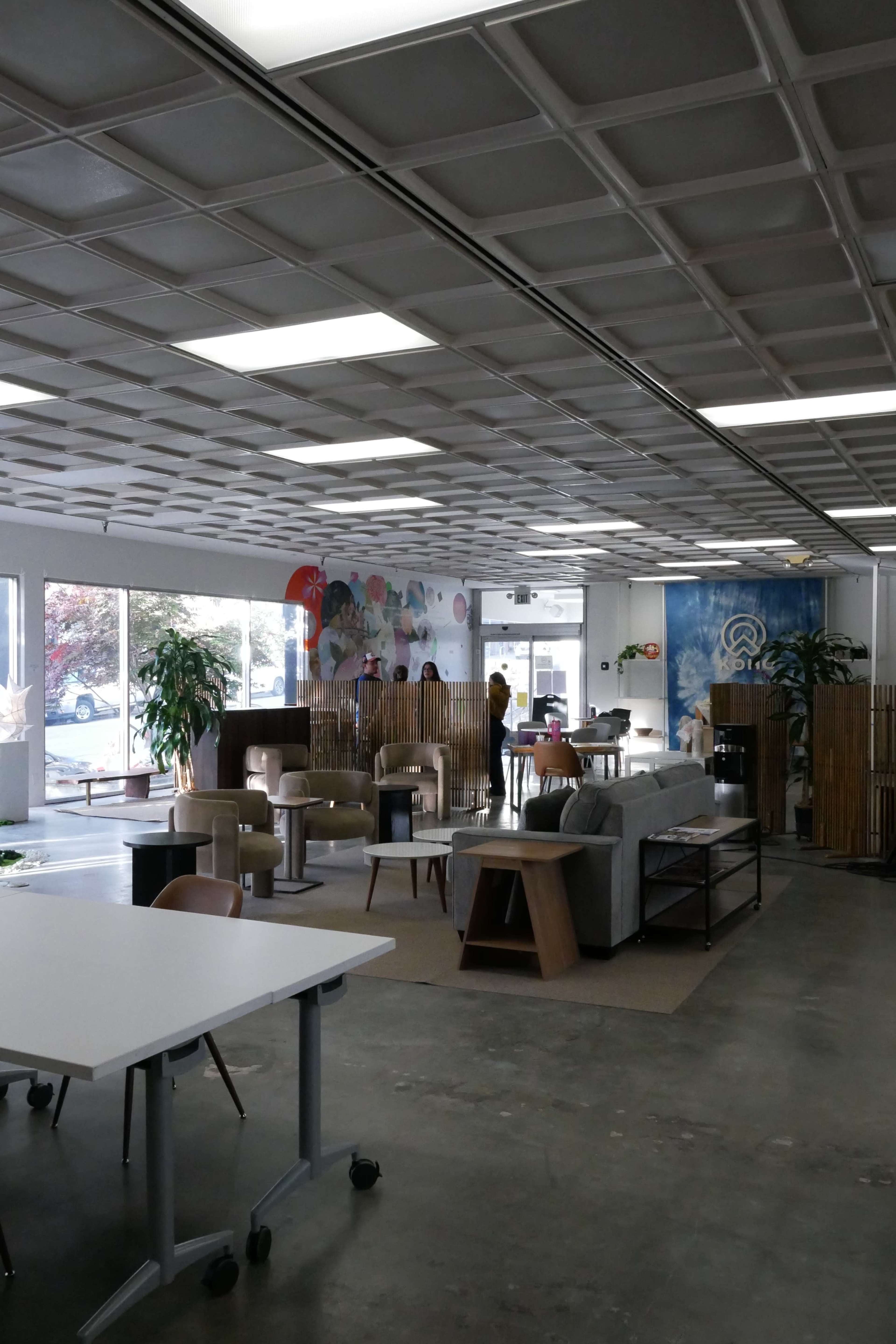 The image shows a spacious, modern interior with various seating areas, tables, and decorative plants, highlighted by a mural on the wall.