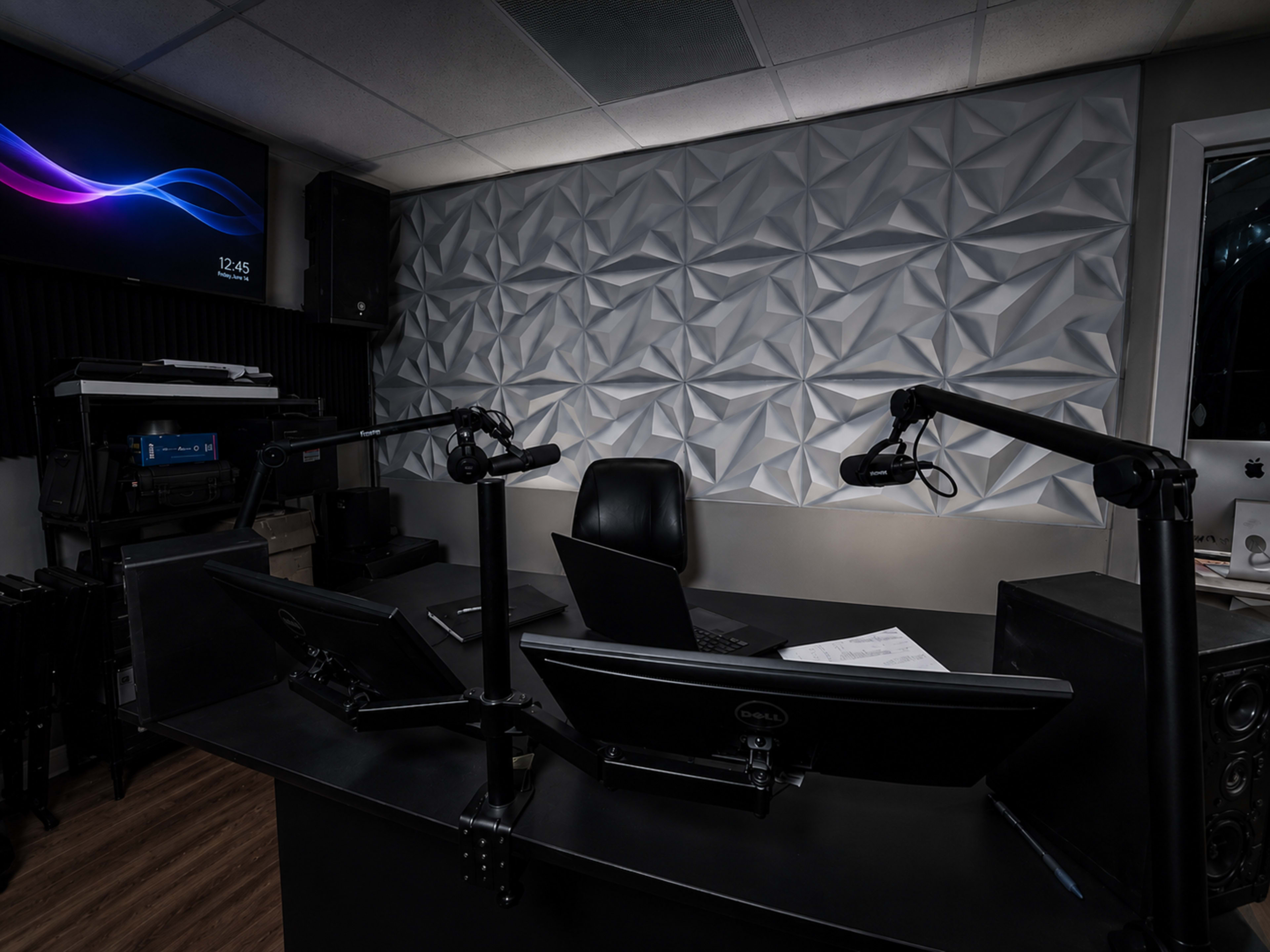 The image shows a modern recording studio with two computer monitors, a black chair, and a textured acoustic wall panel.