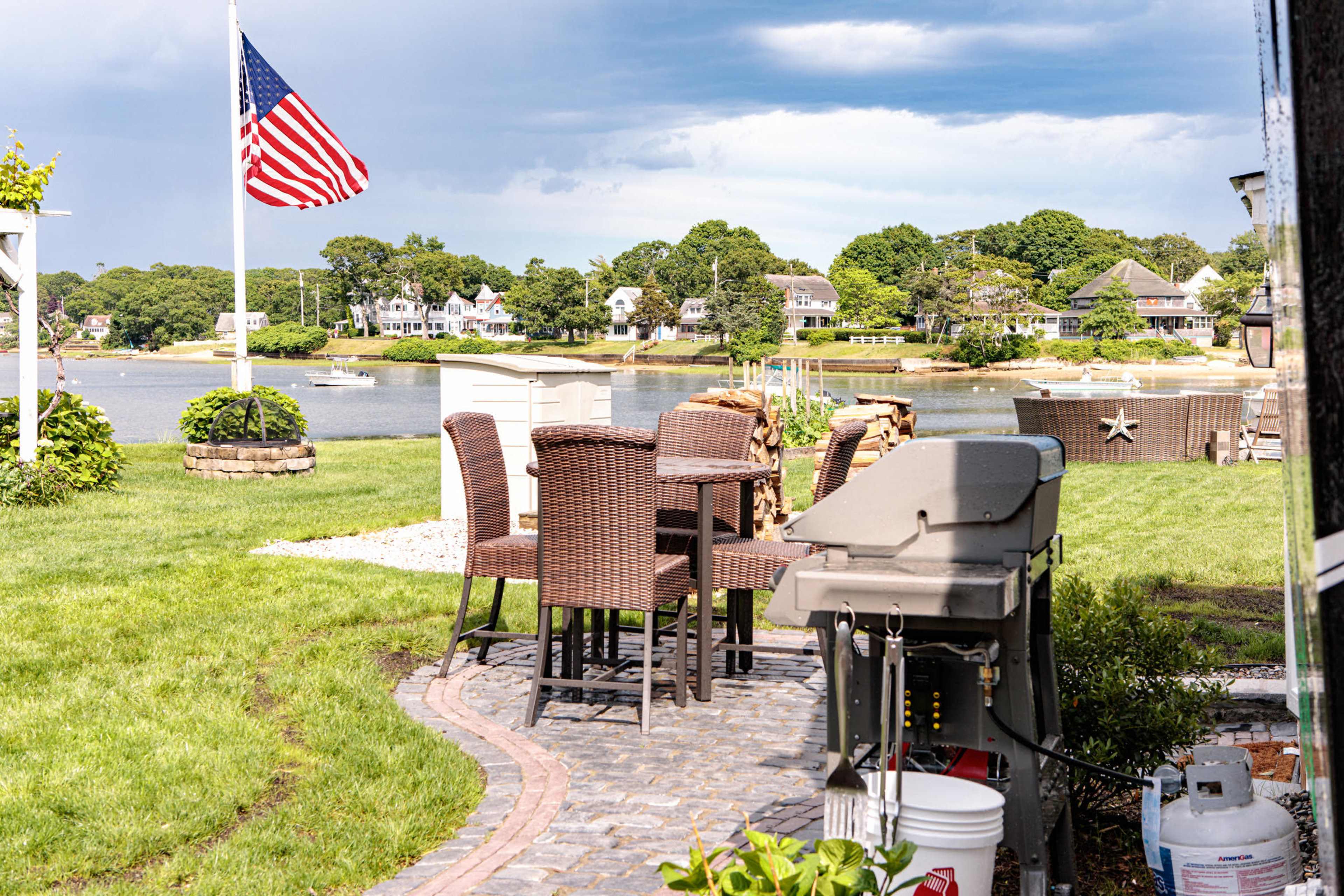 A backyard patio with a grill and seating area, overlooking a body of water and a row of houses across the shoreline.