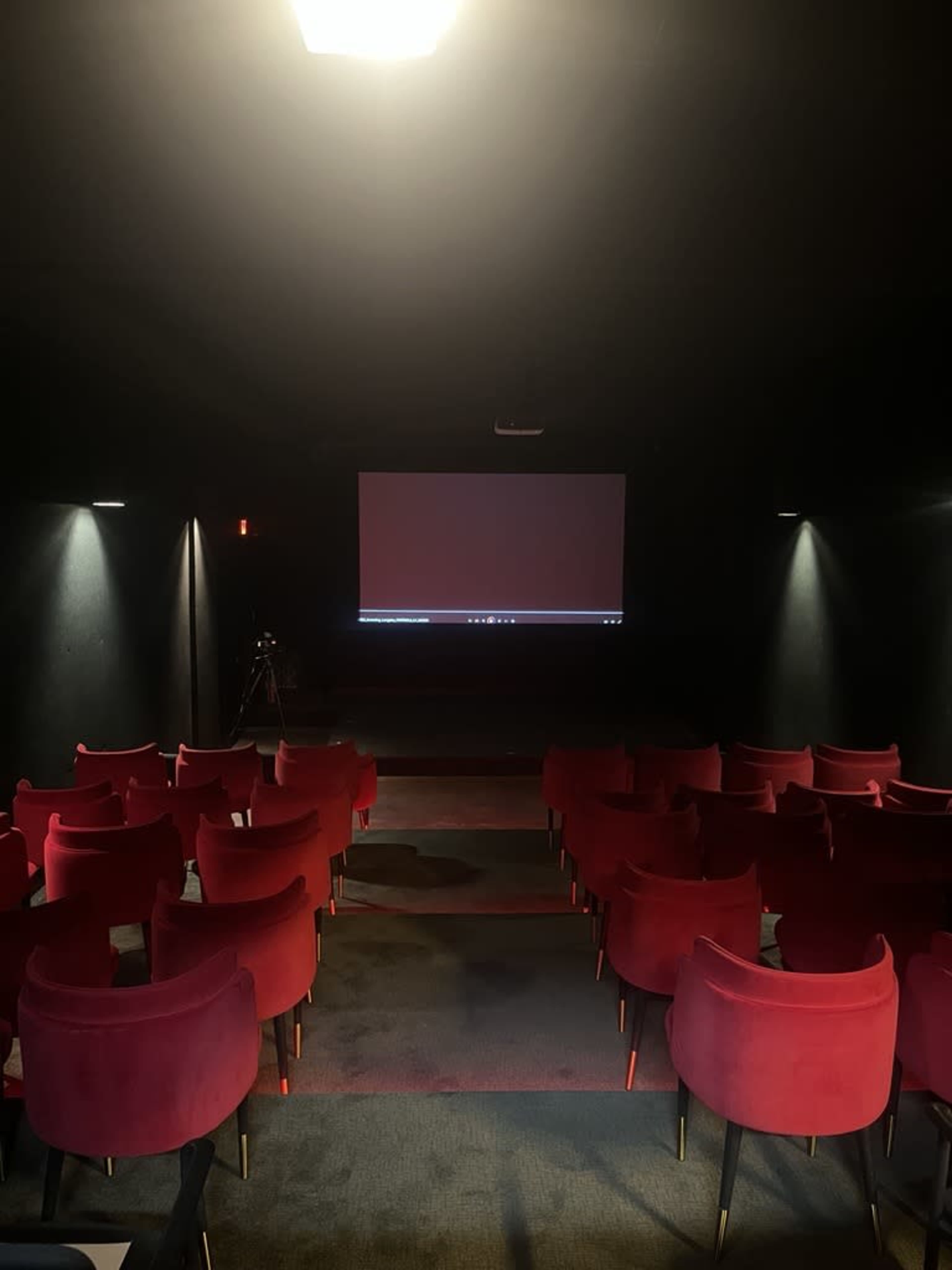 The image shows a darkened theater room with red chairs arranged in rows facing a large screen.