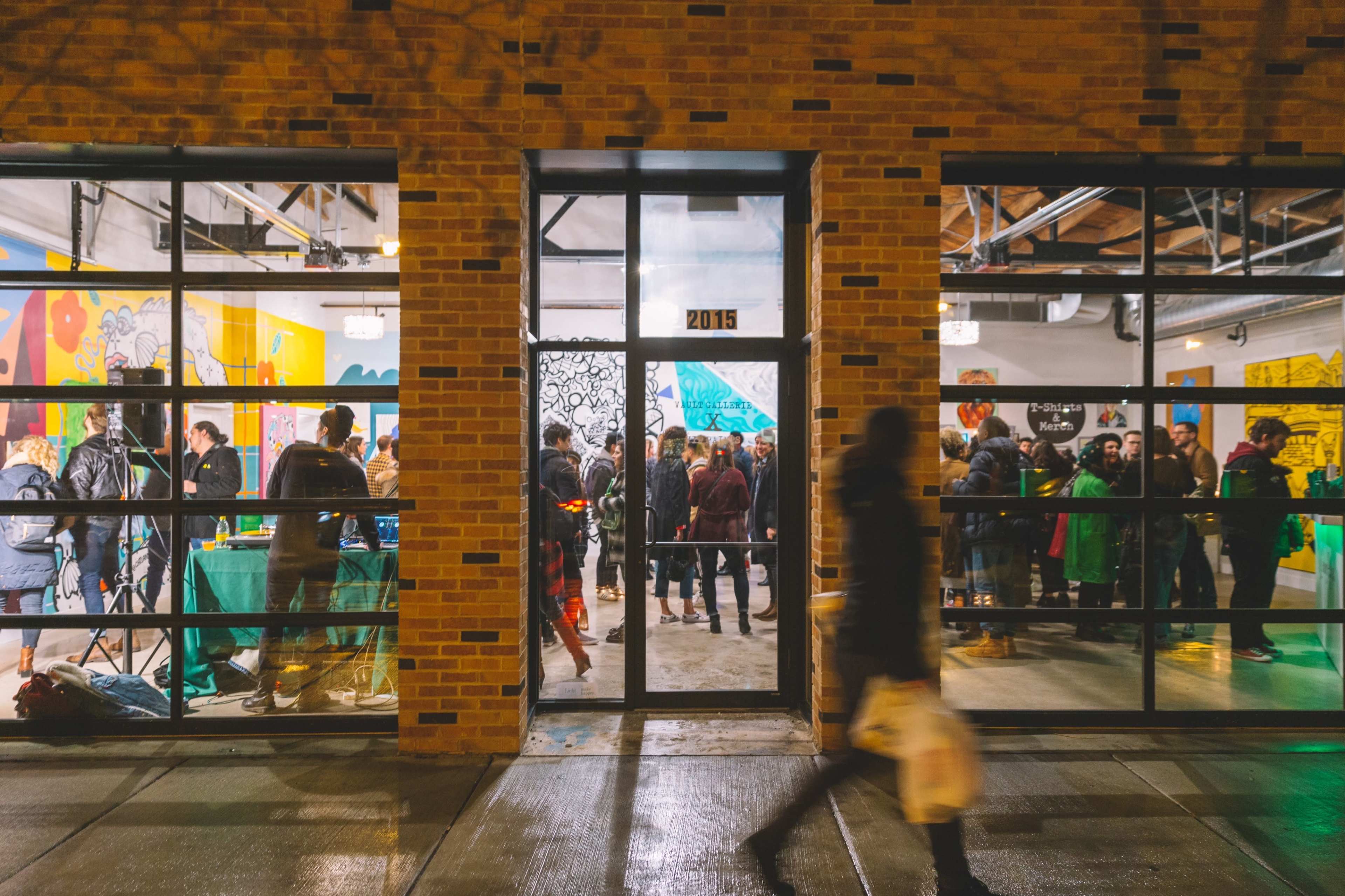 A large crowd gathers inside an art gallery, visible through large glass windows, while a few individuals walk past on the sidewalk.