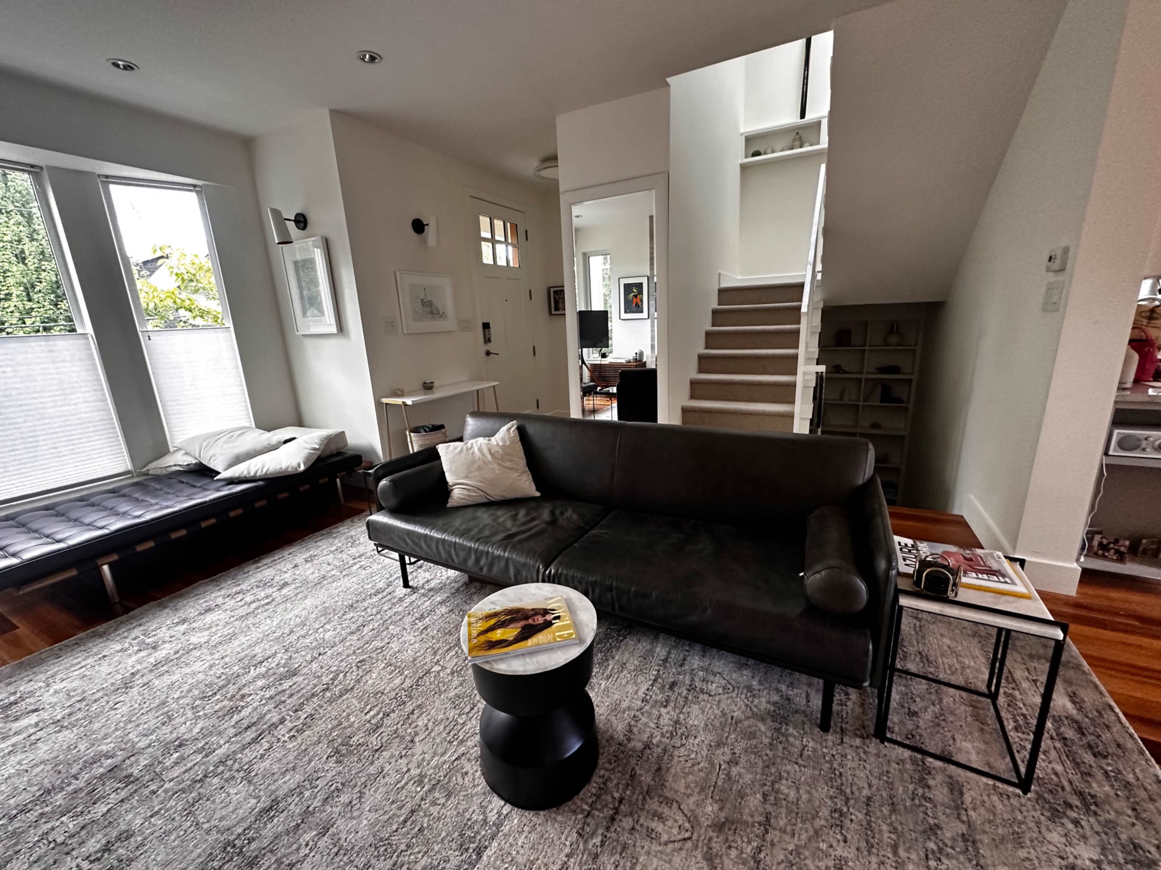 The image shows a modern living room with a black couch, a small side table, and stairs leading to an upper level.