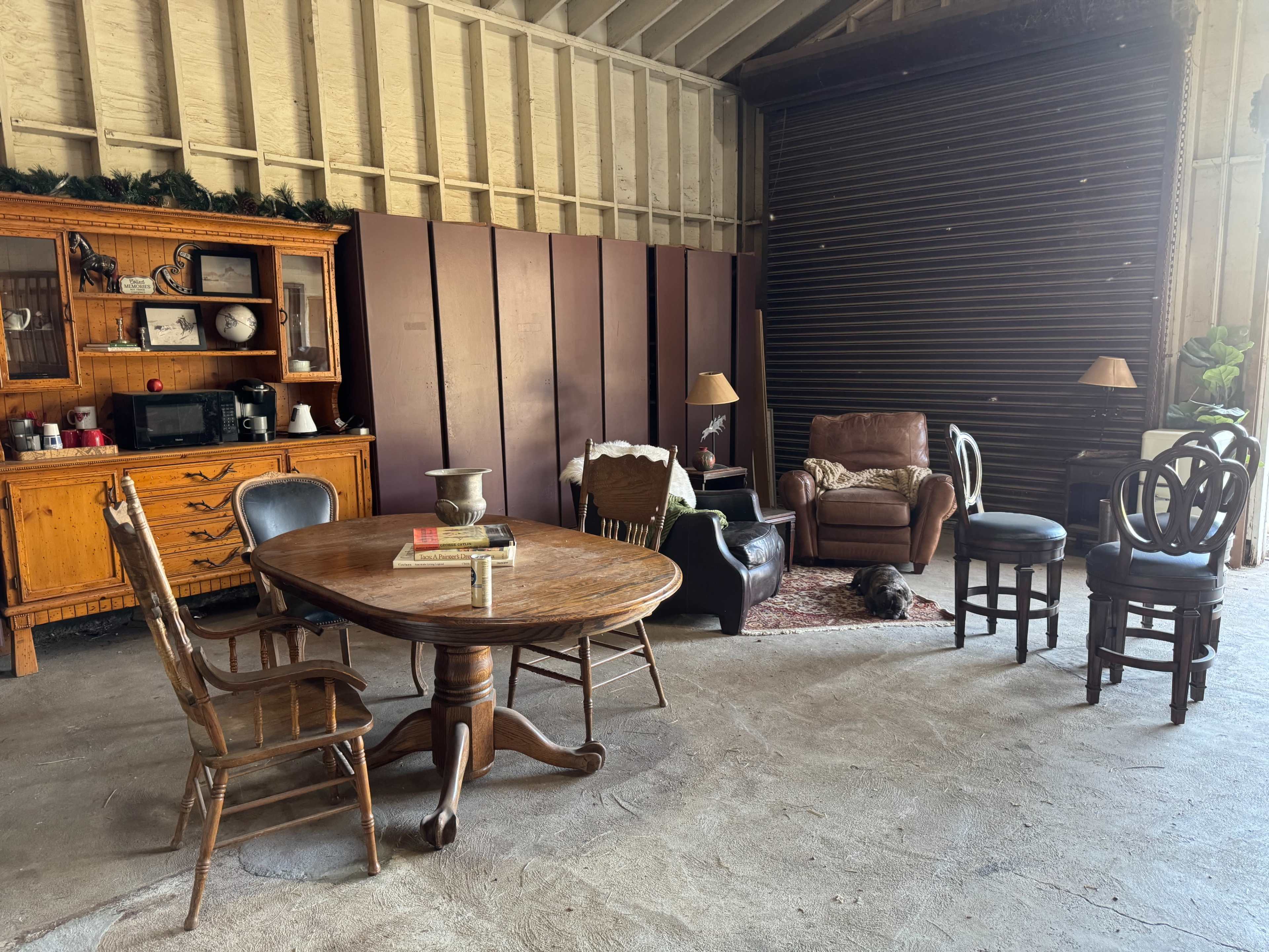 The image shows a spacious, unfinished room with wooden furniture, including a round dining table, several chairs, a couch, and a sideboard against the wall.