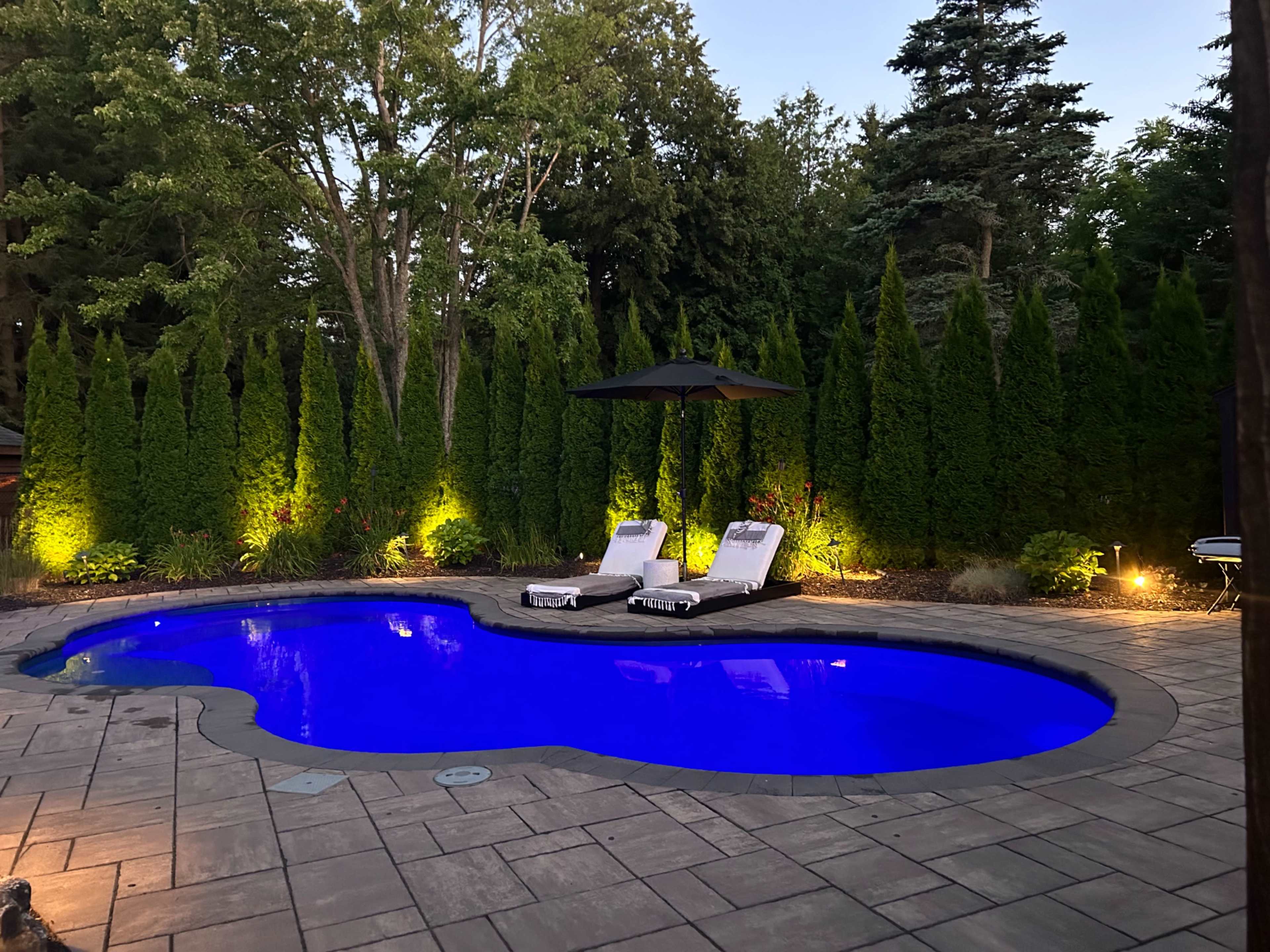 The image shows a swimming pool surrounded by tall green hedges, with two lounge chairs and an umbrella beside the water, illuminated by soft lighting at dusk.