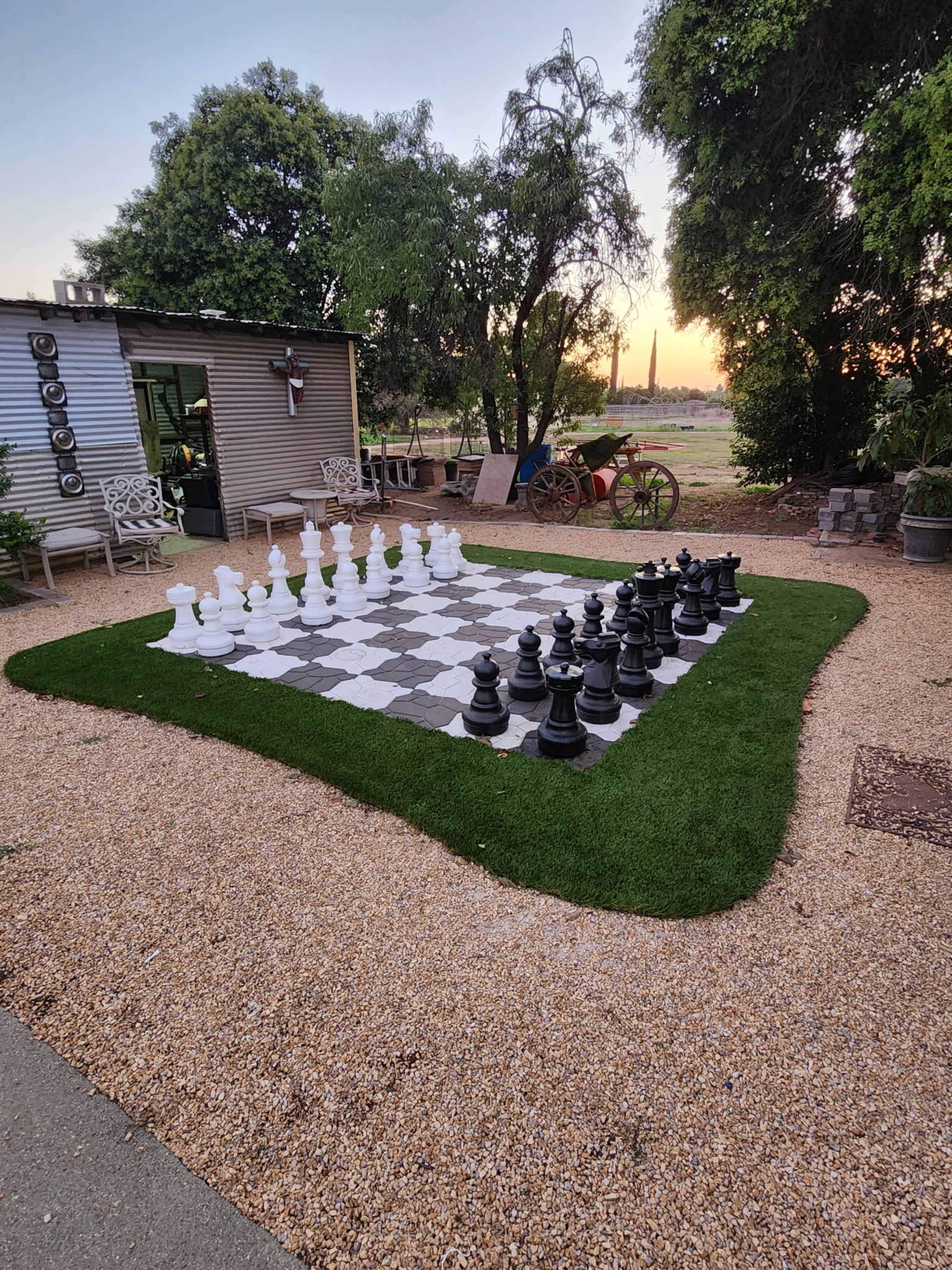 A large outdoor chess board with black and white pieces is set up on a grassy area surrounded by trees and outdoor furniture.