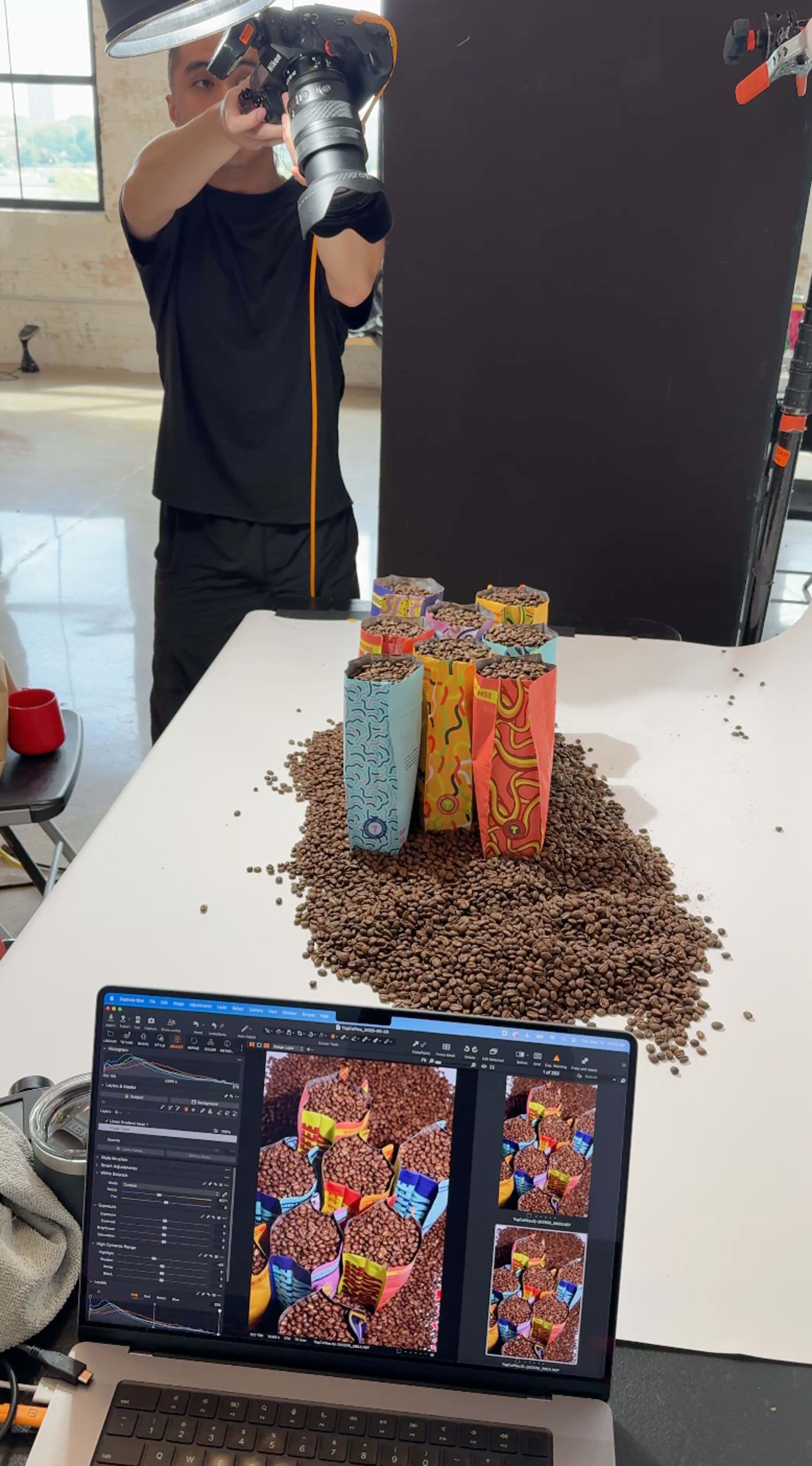 A person stands behind a camera while colorful cartons are arranged on a mound of coffee beans on a table.