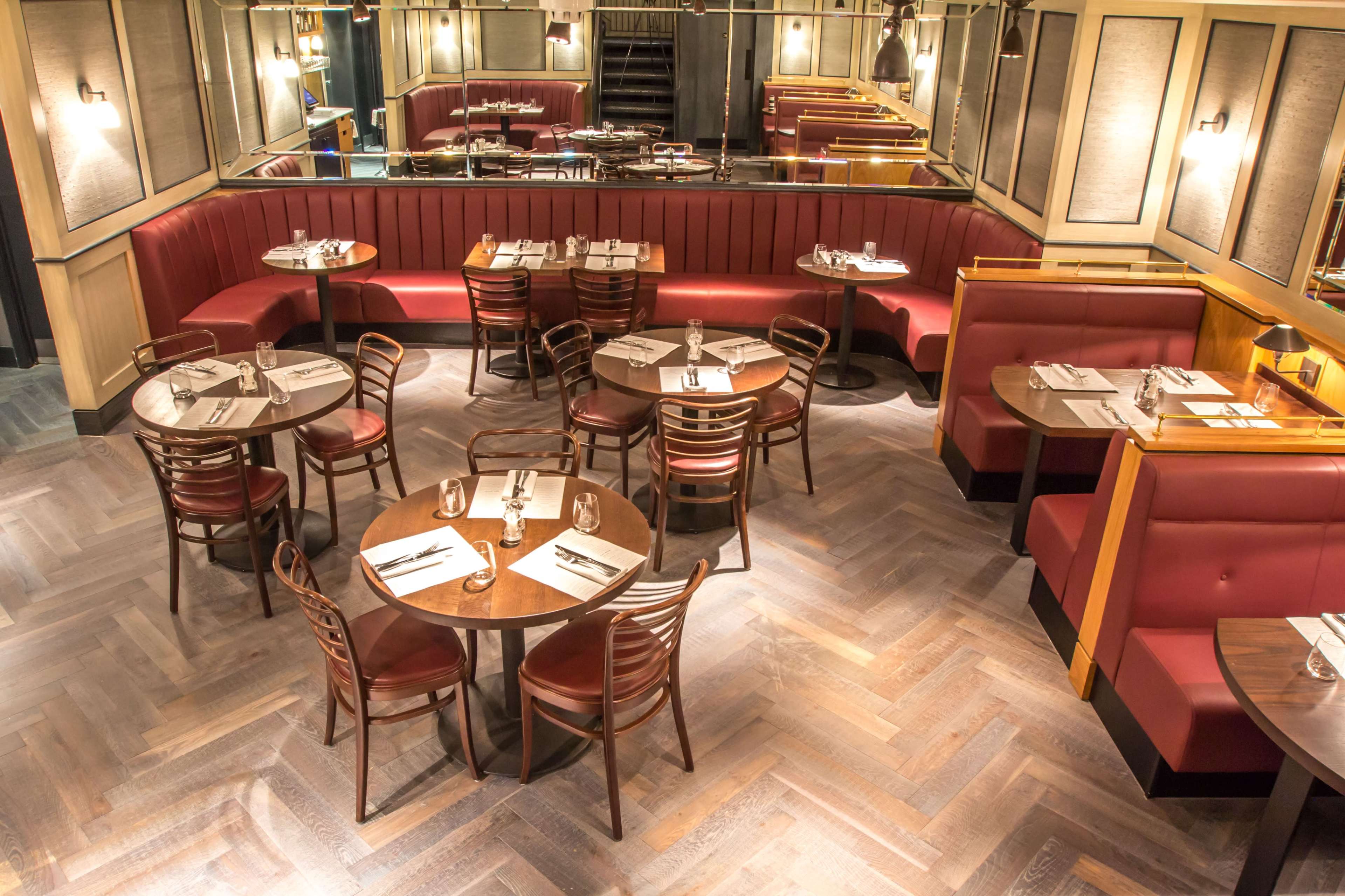The image shows a restaurant interior featuring wooden tables surrounded by chairs and banquettes, with a mirrored wall reflecting the seating arrangement.
