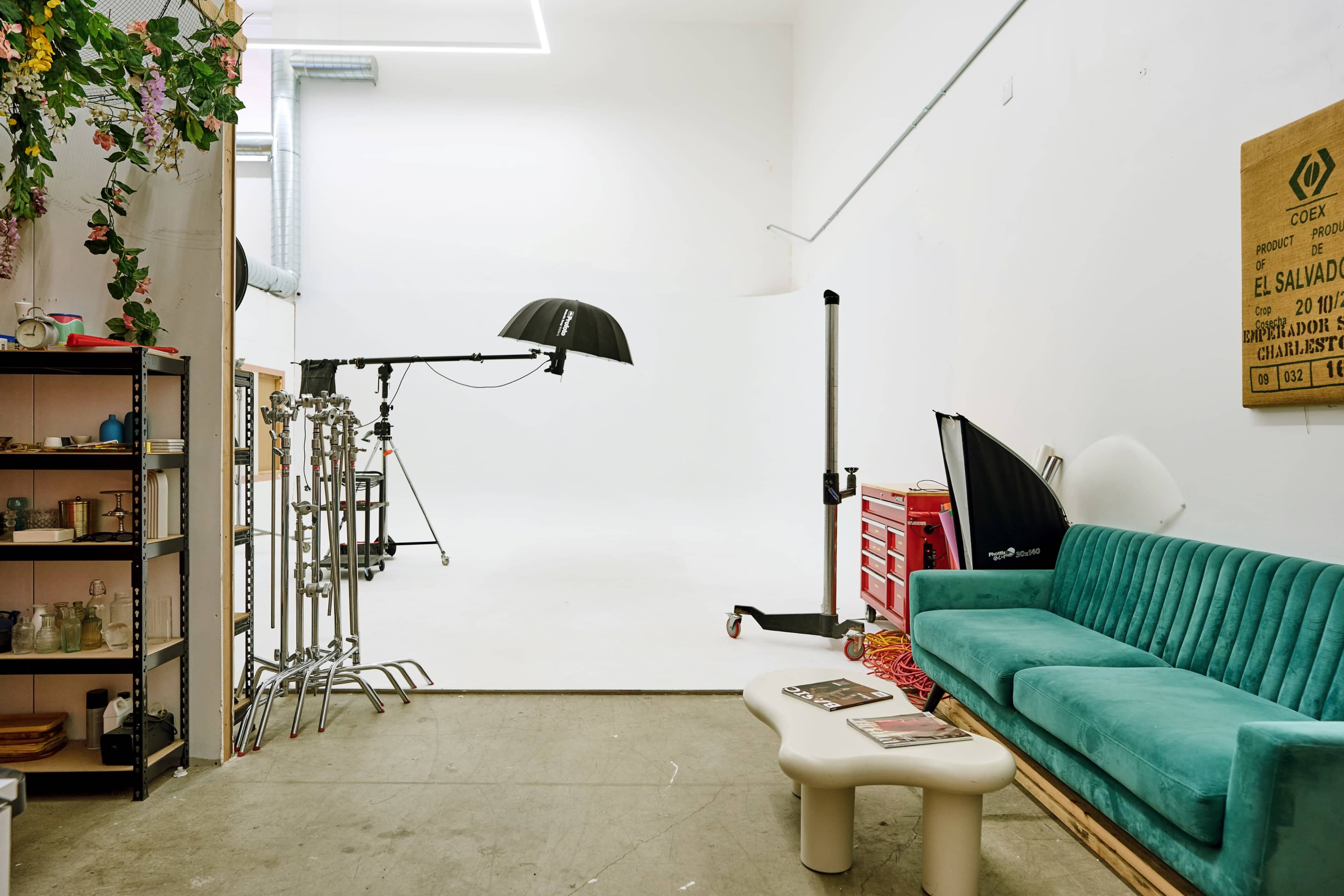 The image shows a studio space featuring a green couch, a photography light setup, and a shelf with various items, all against a white backdrop.