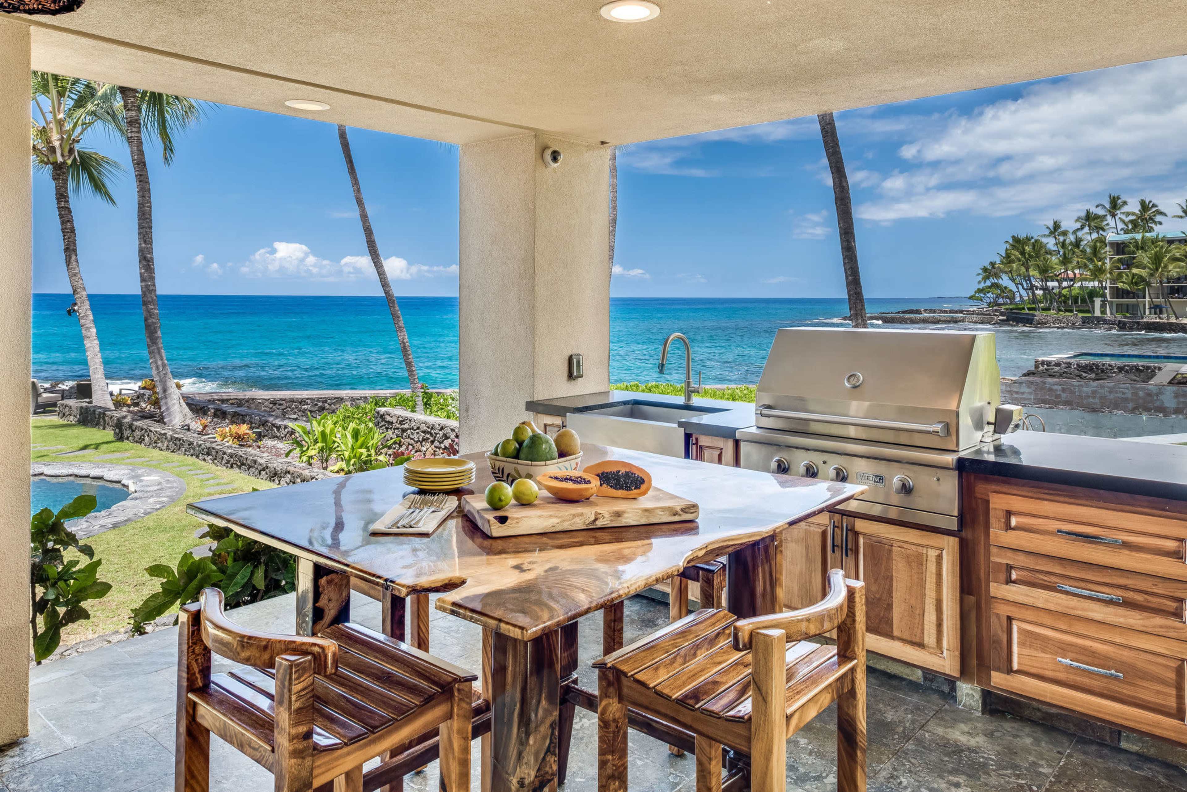 An outdoor kitchen with a barbecue grill and a dining table overlooks the ocean, framed by palm trees and a clear blue sky.