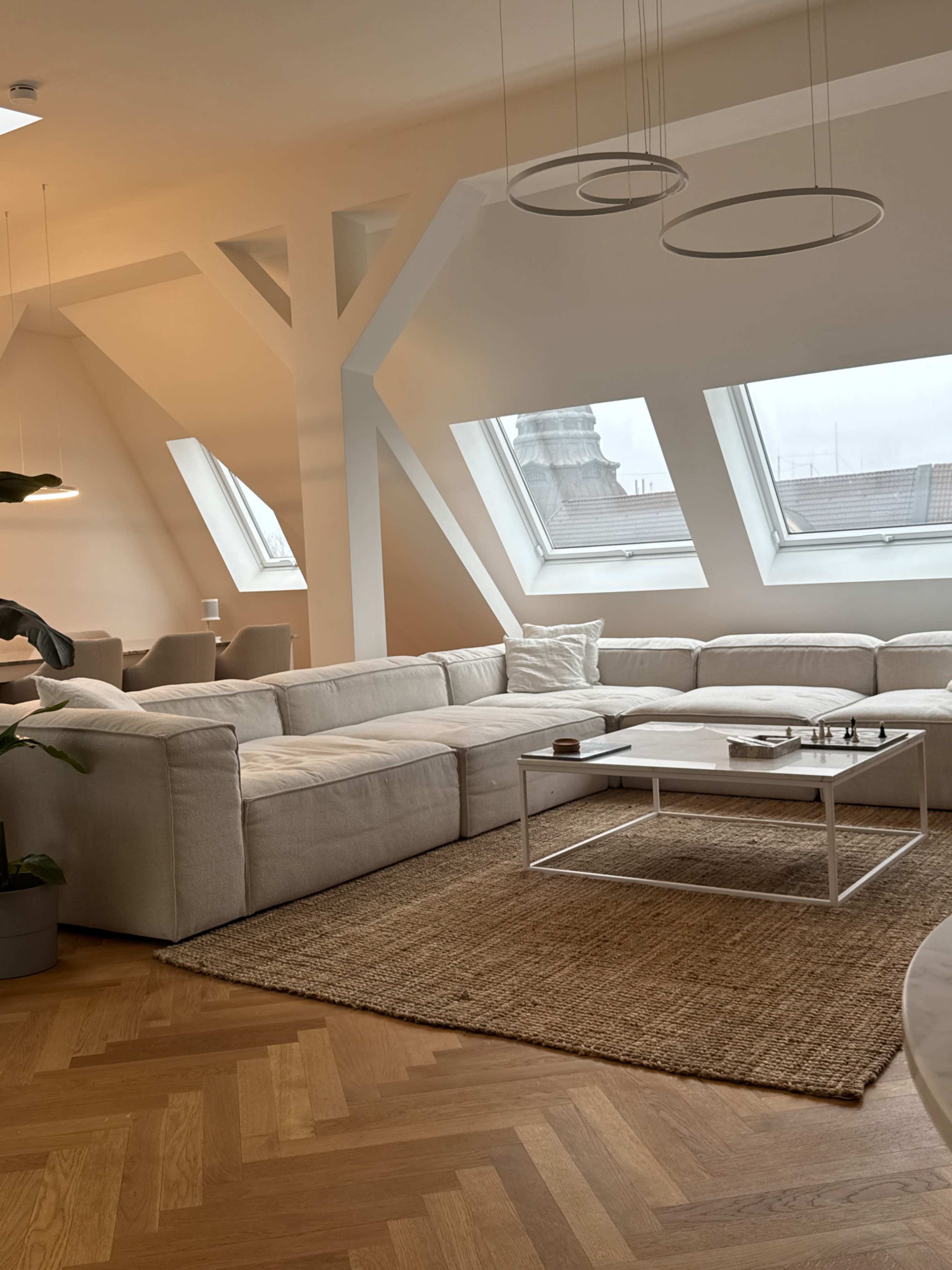The image shows a spacious, modern living room with a large sectional sofa, wooden flooring, and several skylight windows.