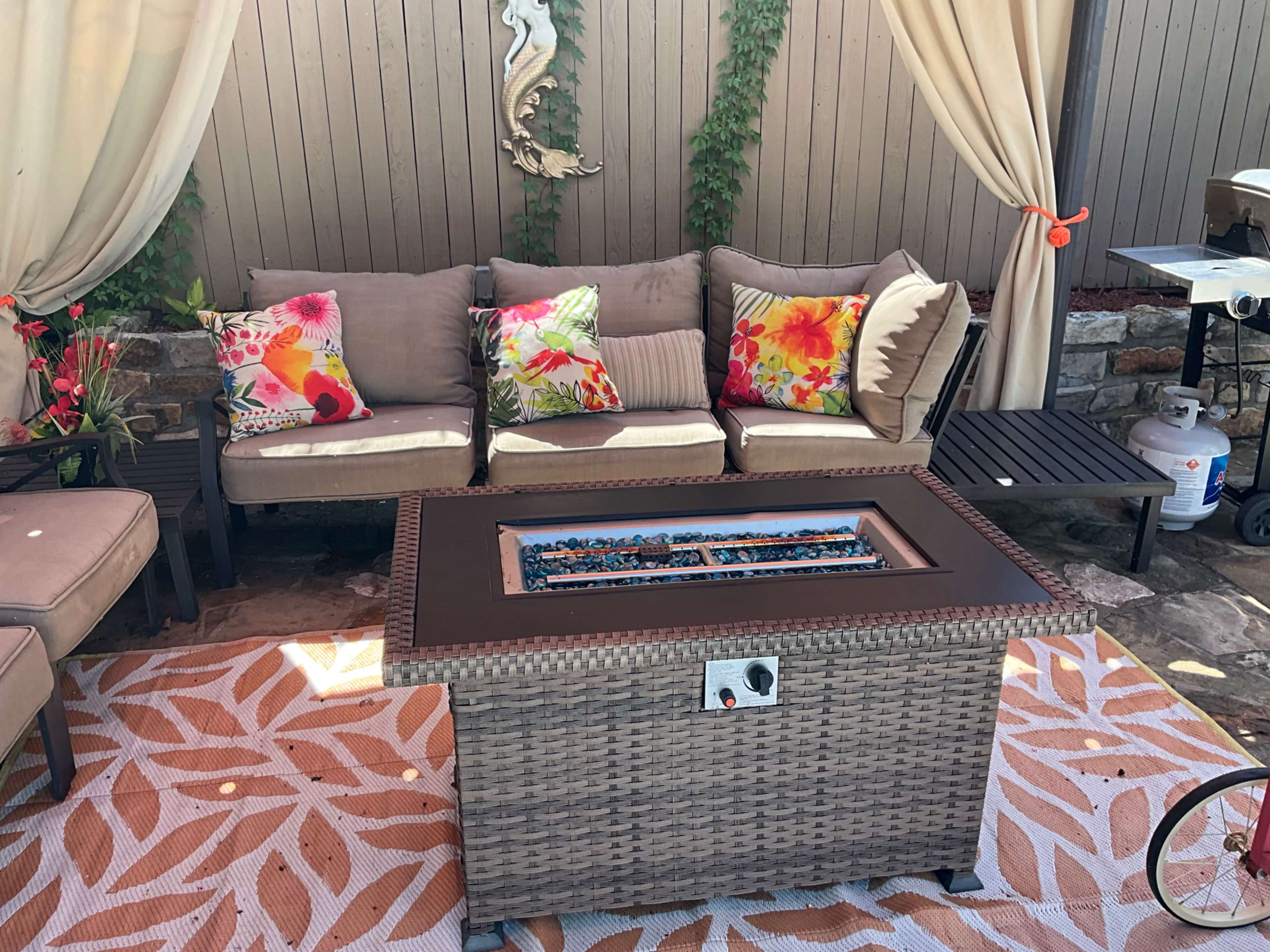 The image shows a cozy outdoor seating area with a woven fire pit table surrounded by cushions on a sectional sofa and a patterned rug.