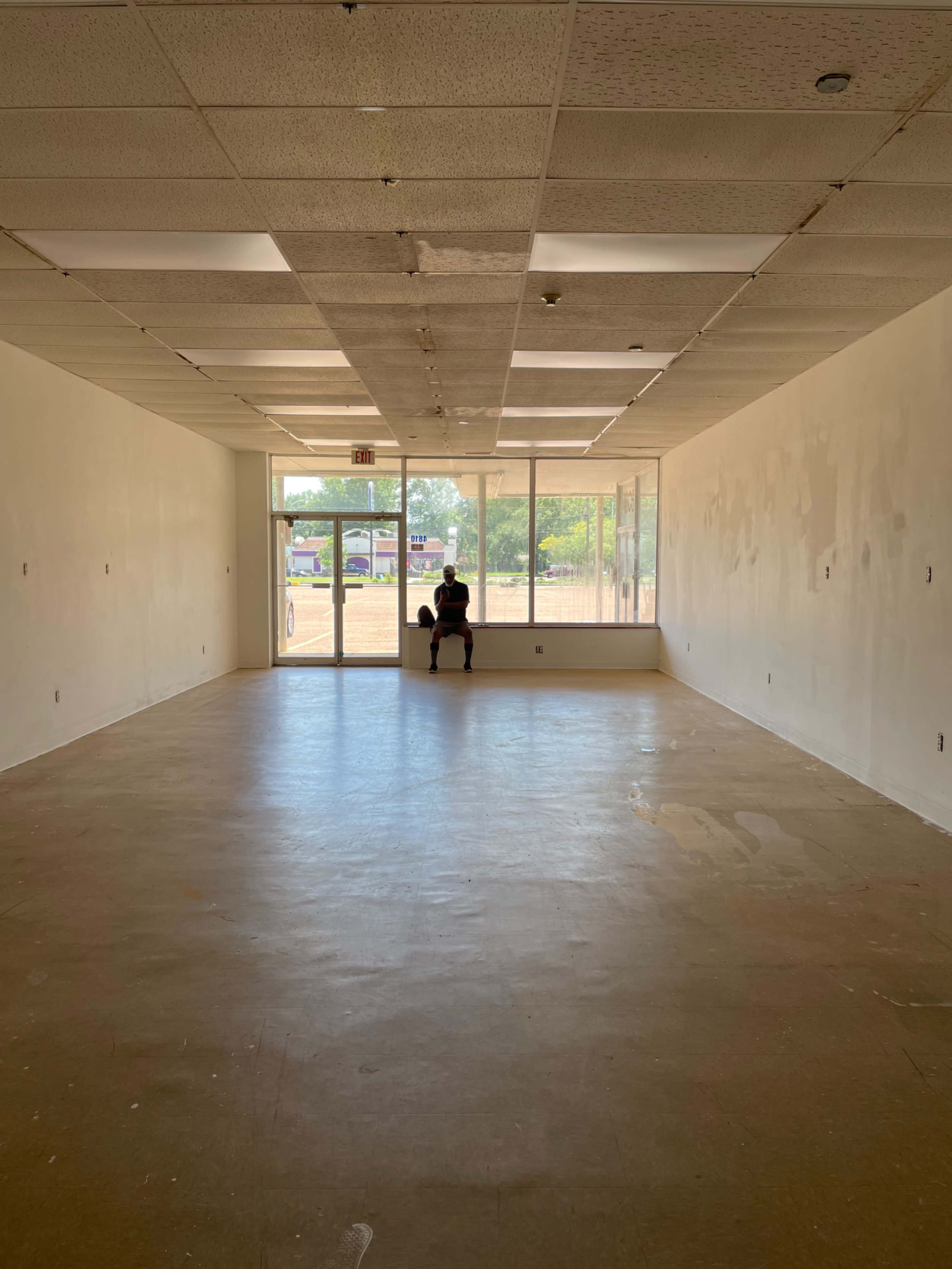A person sits alone in a vacant room with large windows facing the entrance.