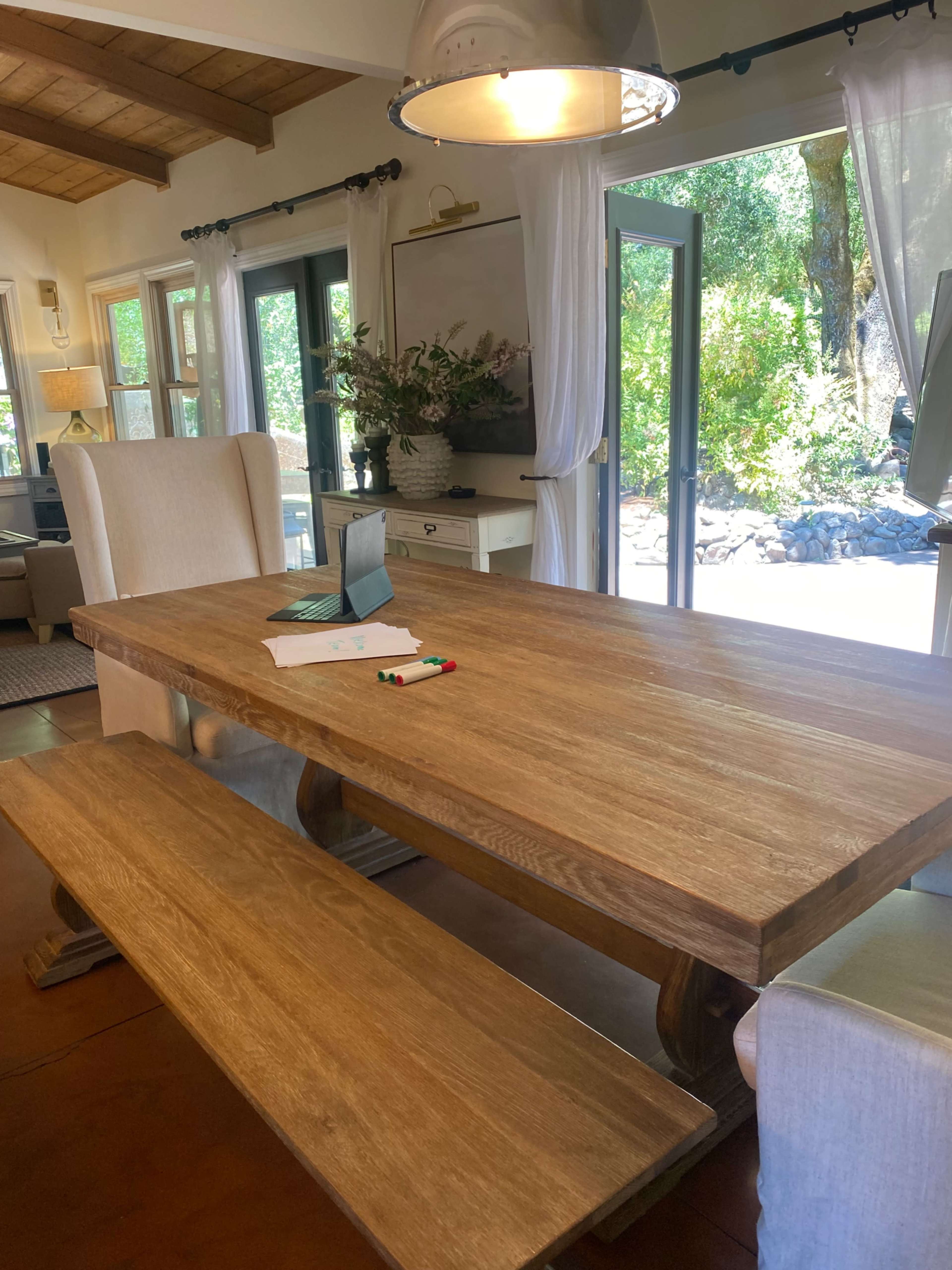 A wooden dining table with a bench is placed in a light-filled room featuring large windows and greenery visible outside.