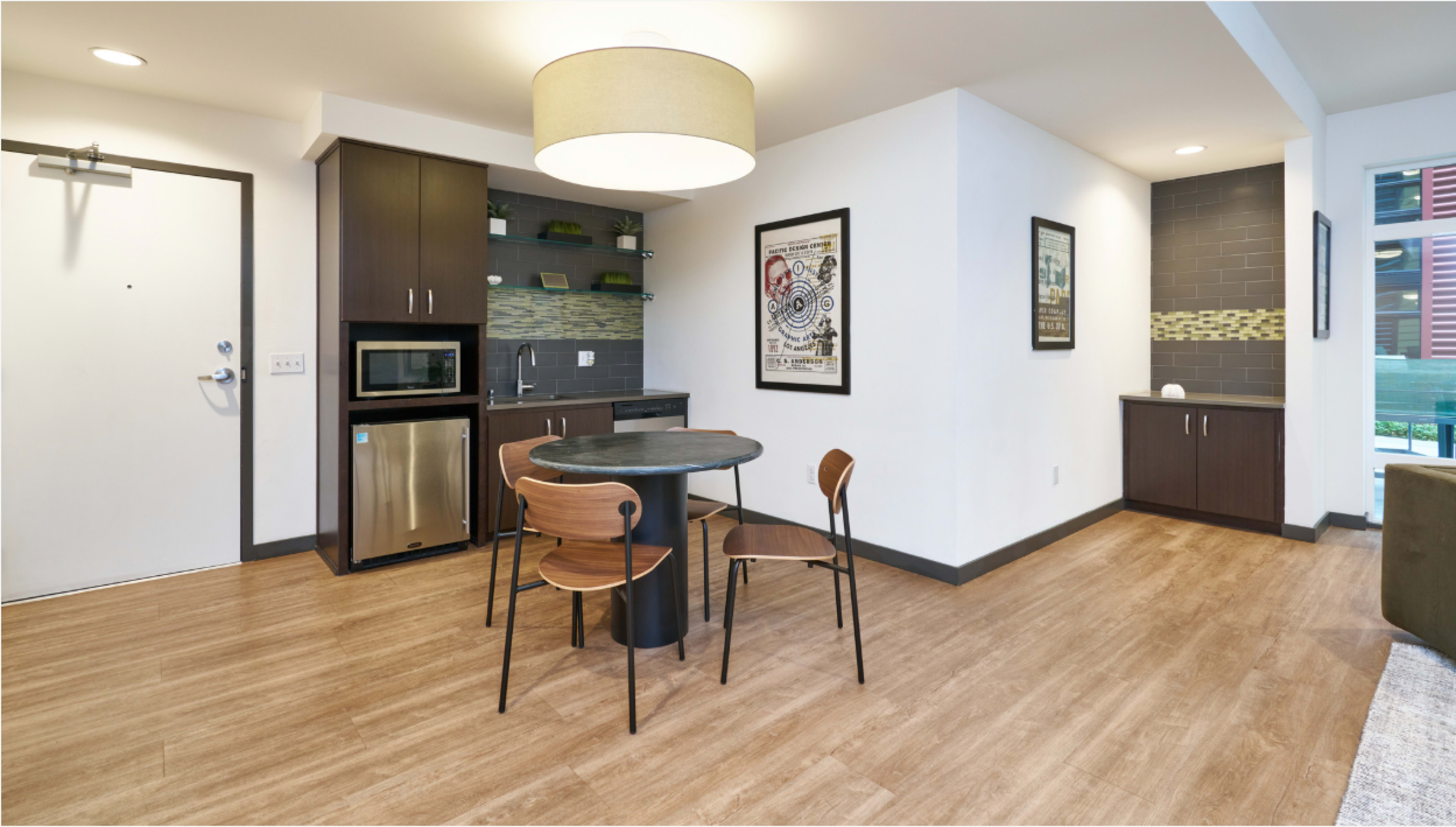 The image shows a modern kitchen-dining area with a round table and two chairs, a kitchenette with a microwave and fridge, and a decorative wall featuring framed artwork.