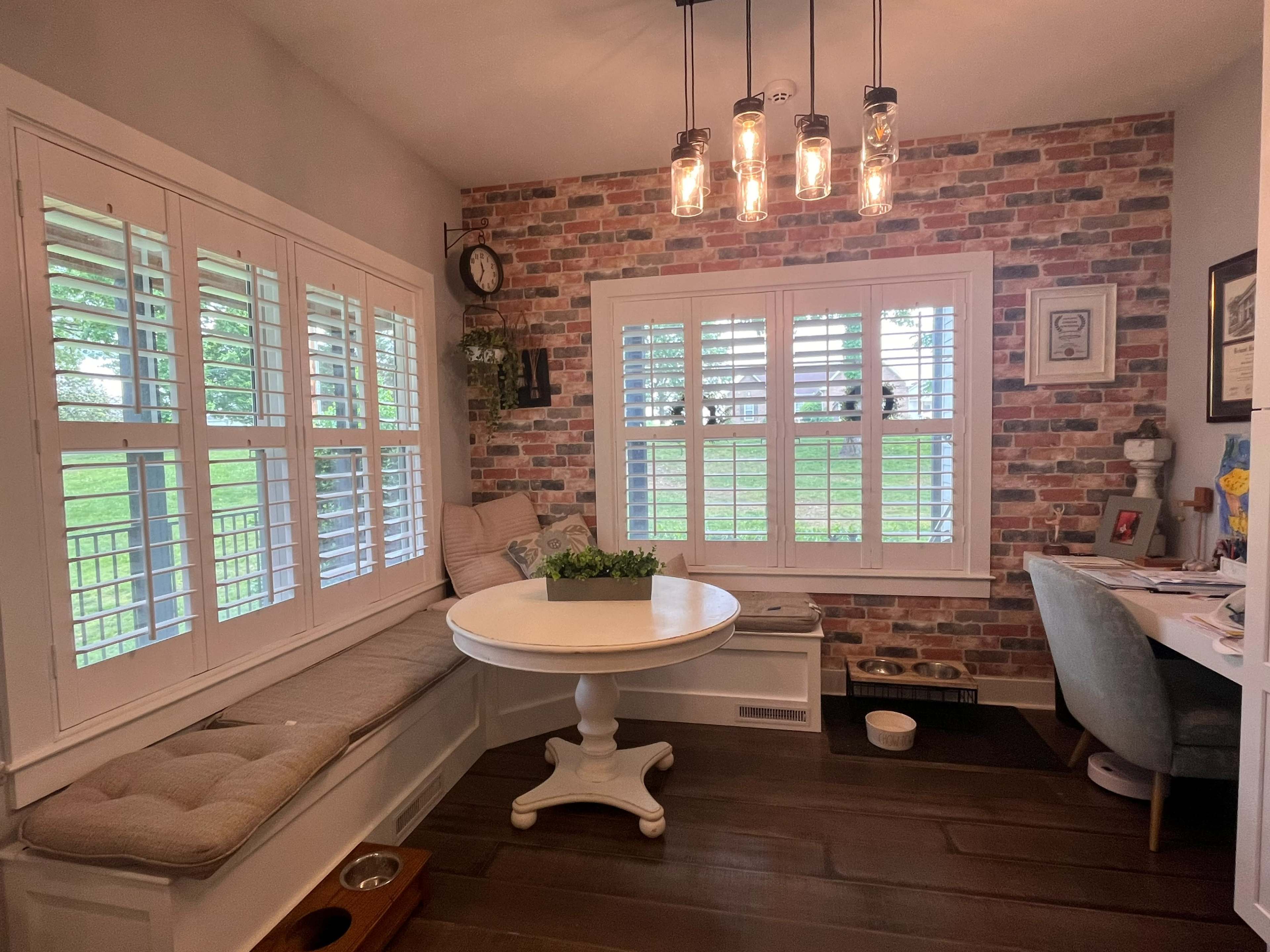 The image shows a cozy nook with a round table, a built-in bench seat with cushions, and a desk in a room featuring brick-patterned wallpaper and plantation shutters.