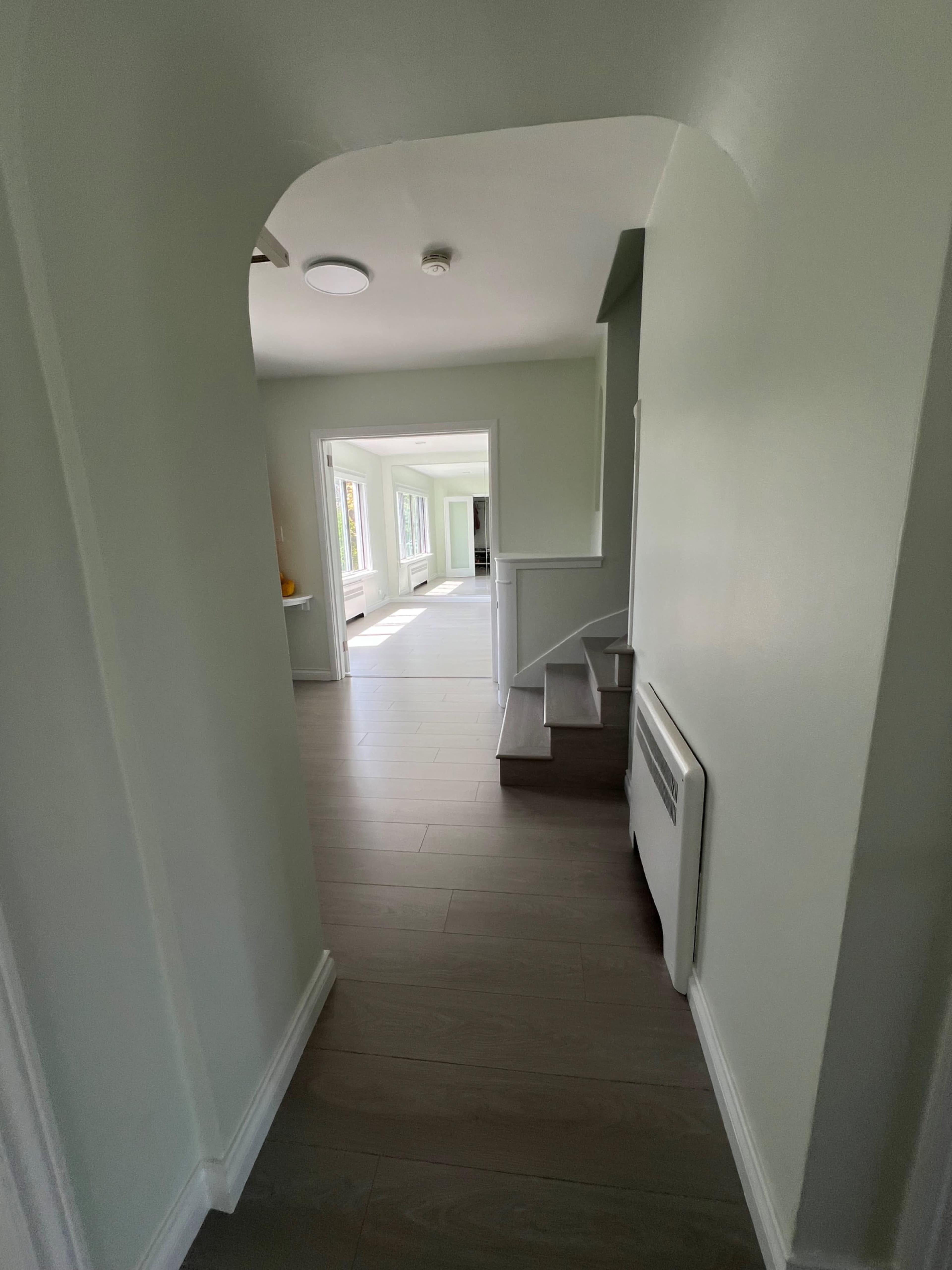 A hallway leads to a sunlit room with large windows, featuring a staircase on the right and a smooth floor.