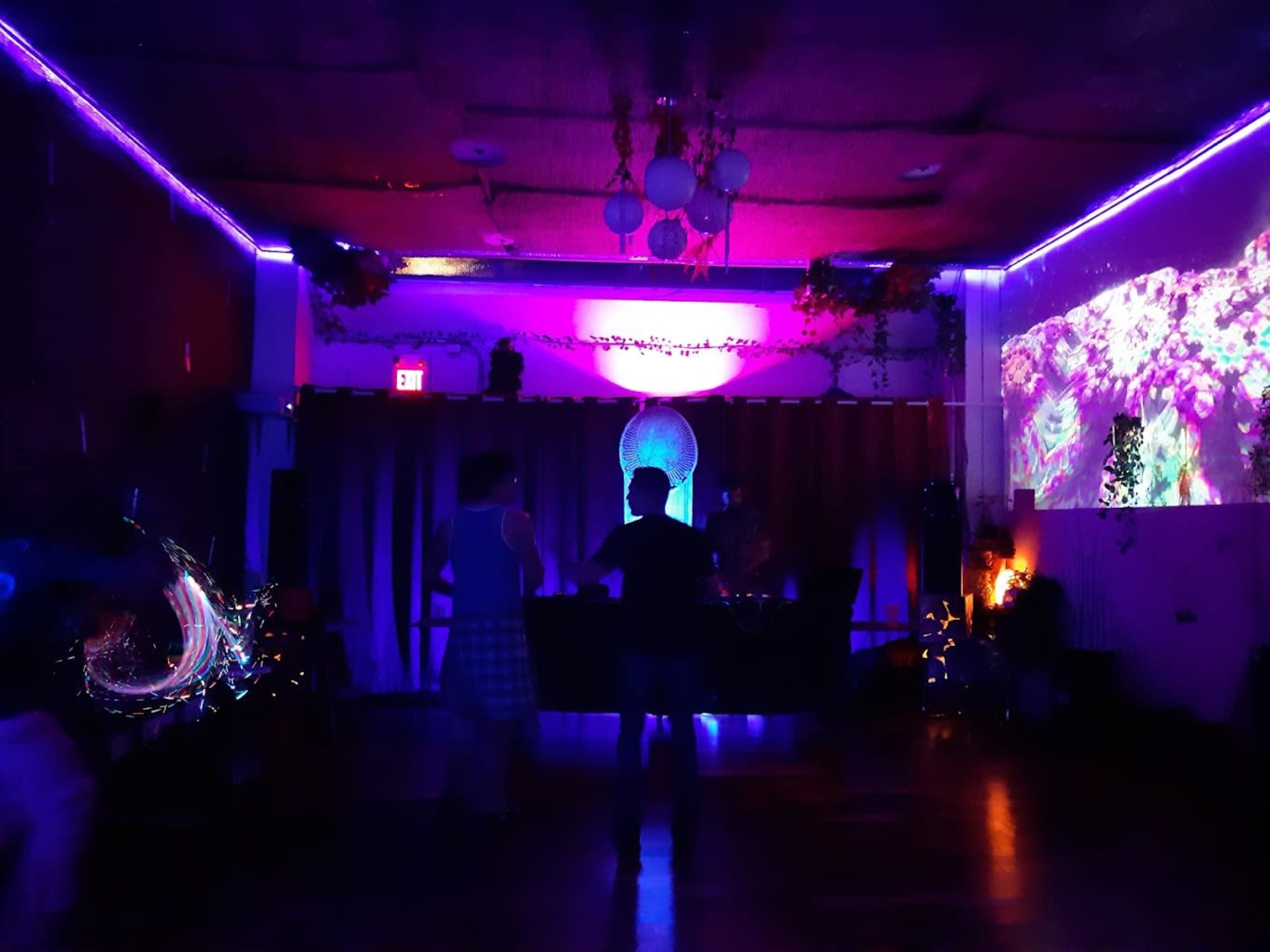 A small, dimly lit dance hall features purple neon lights, with a DJ booth at the front and people dancing on the floor.