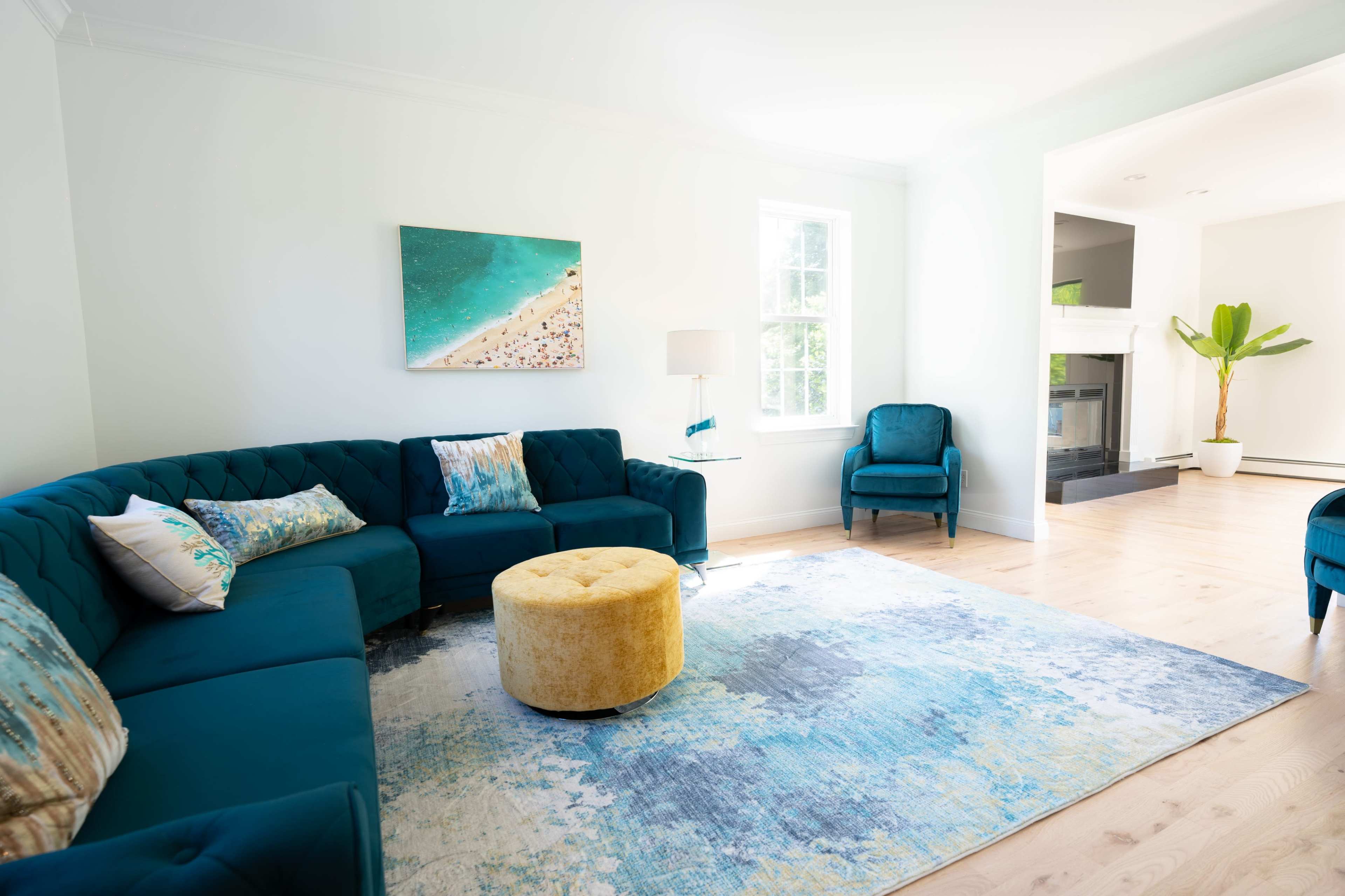 A bright living room features a blue sectional sofa, a round yellow ottoman, and a light area rug, with a beach-themed painting on the wall.