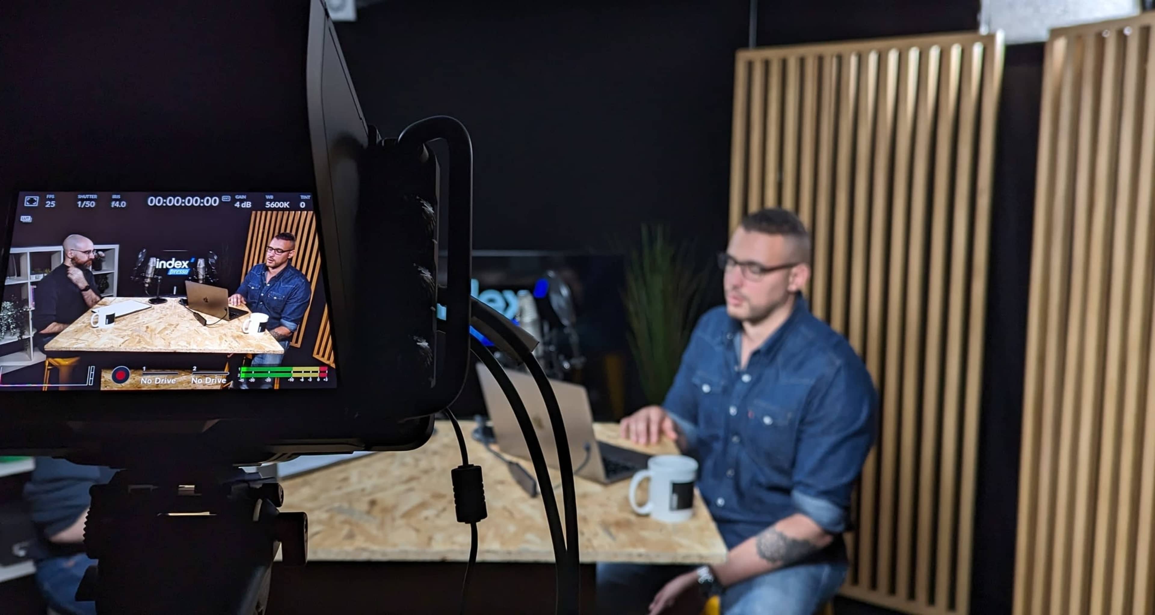 A video recording setup shows a man in a denim shirt seated at a table with a coffee mug, while another man is partially visible on a screen in front of a backdrop with wooden panels.