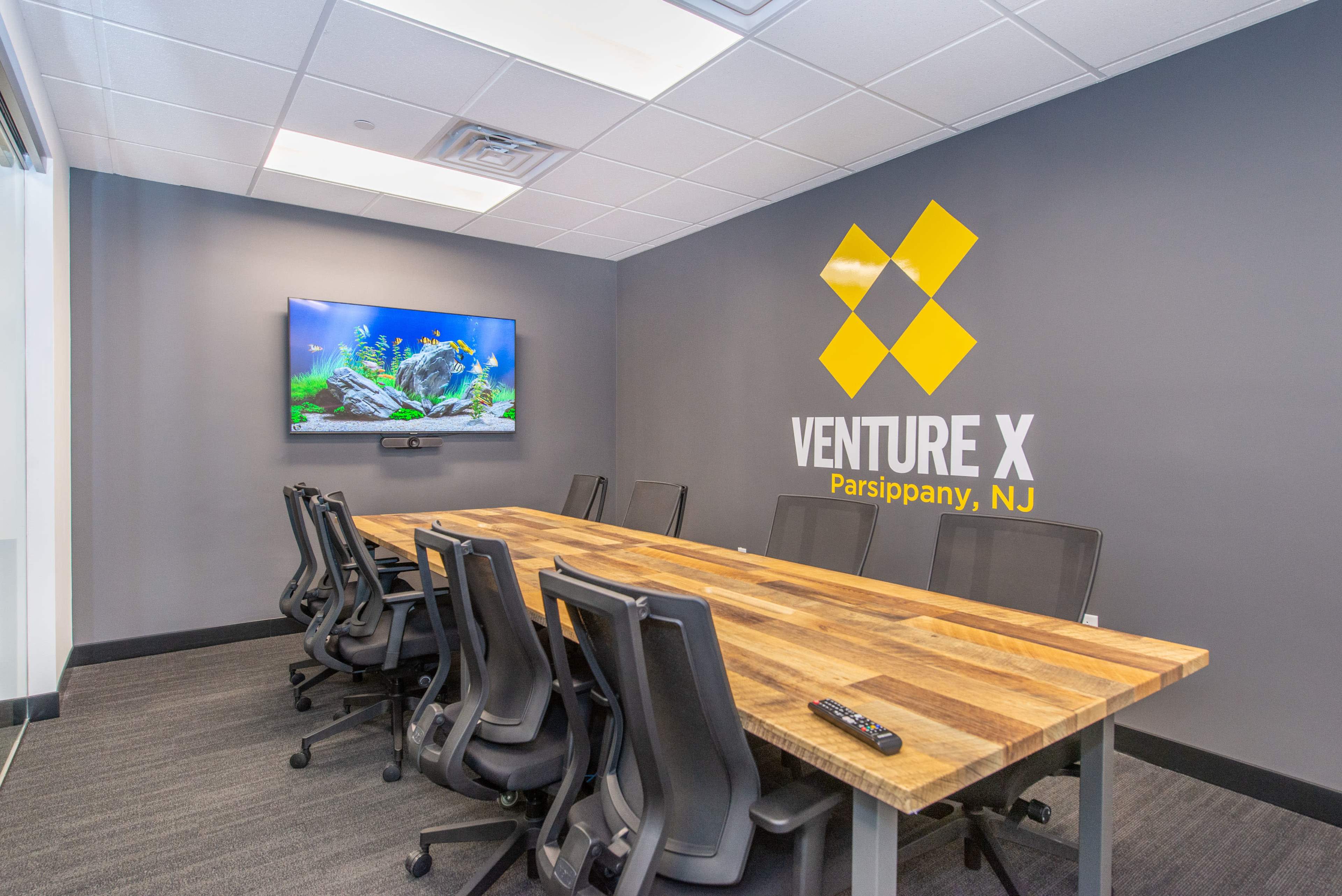 A meeting room with a large wooden table, several black chairs, a wall-mounted TV displaying aquarium visuals, and the "Venture X Parsippany, NJ" logo on the wall.