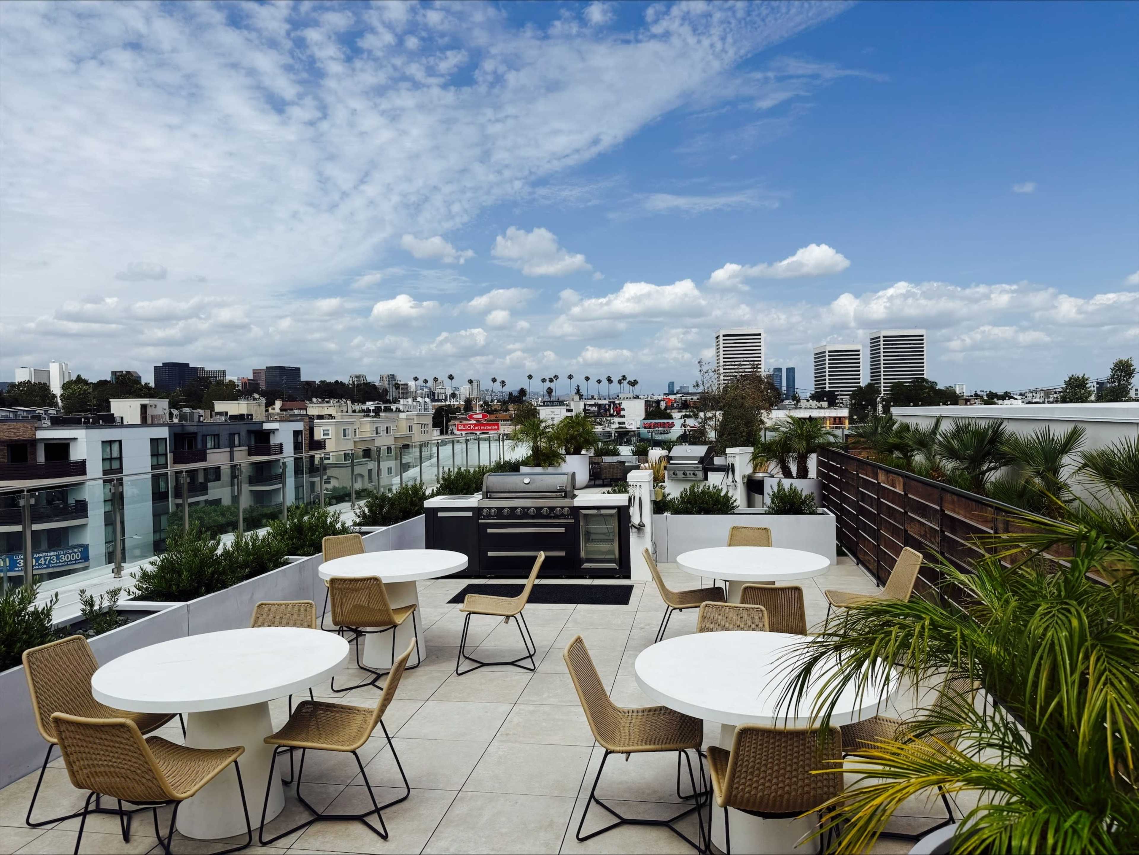 The image shows a rooftop patio with several circular tables and chairs, surrounded by greenery and overlooking a city skyline with low-rise buildings and palm trees under a partly cloudy sky.