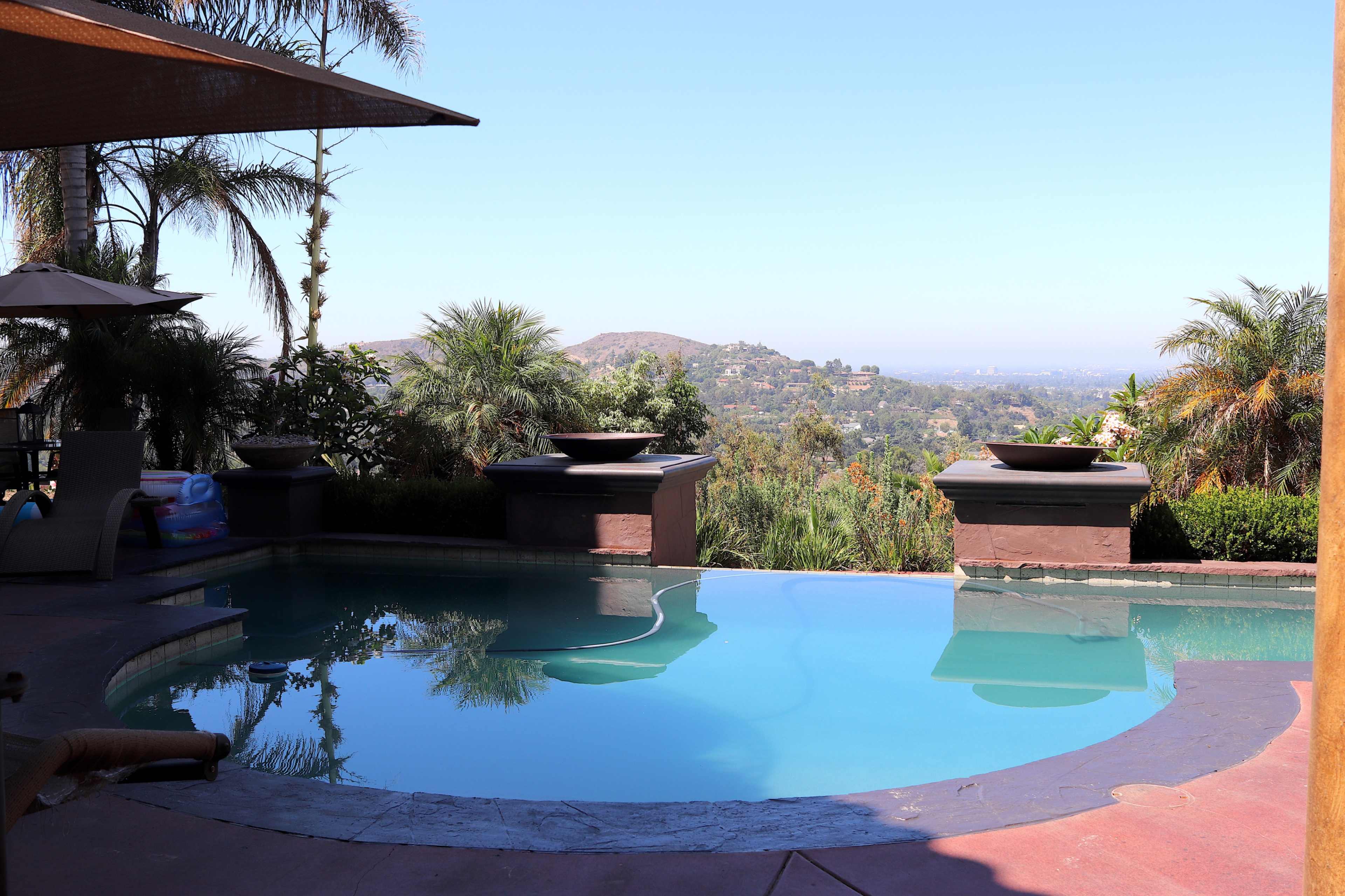 A swimming pool surrounded by palm trees and planters, with a view of rolling hills in the background under a clear blue sky.