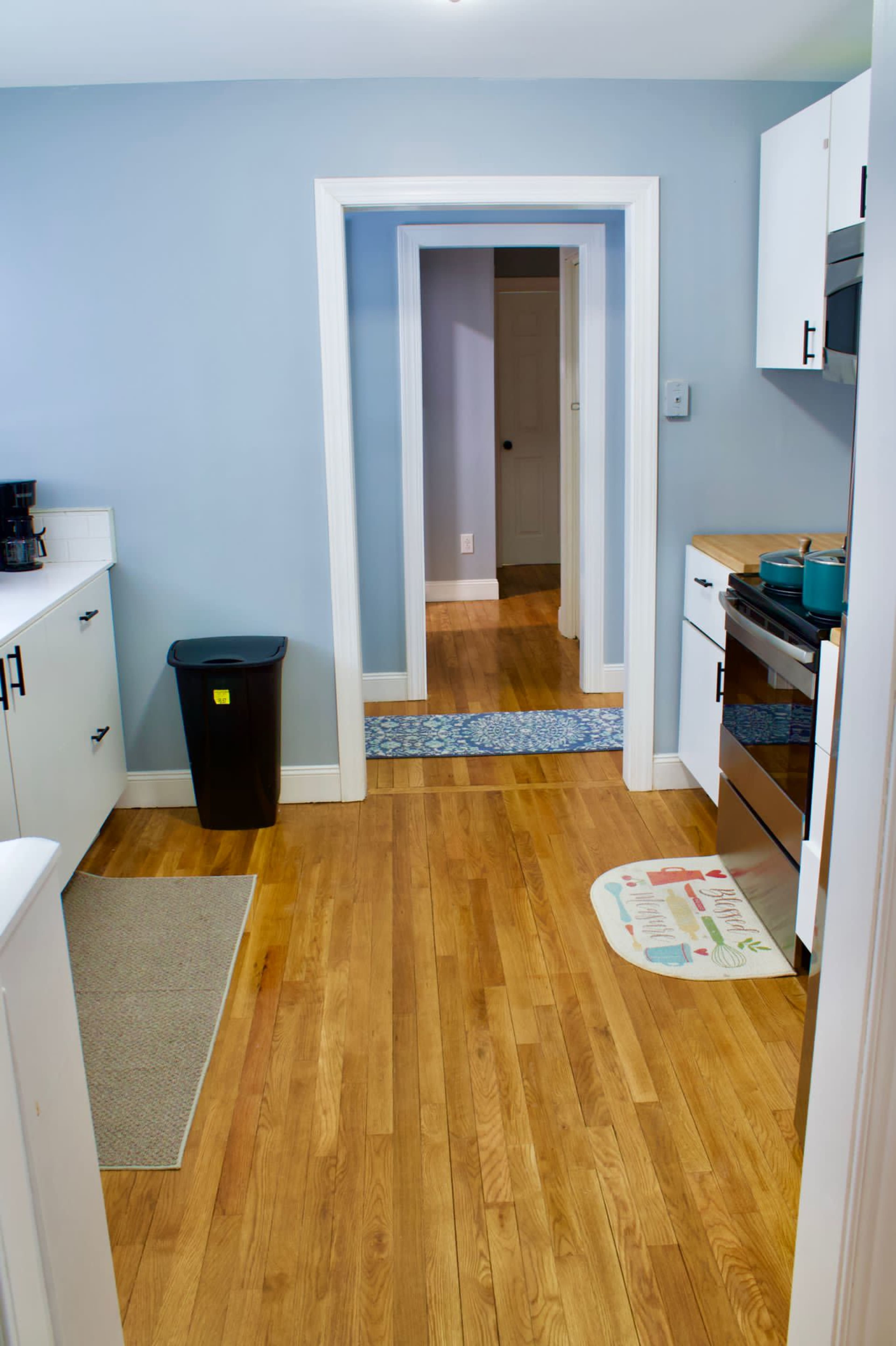 A kitchen with wooden floors, blue walls, and a doorway leading to another room is shown, featuring a black trash can, a gray rug, and stainless steel appliances.