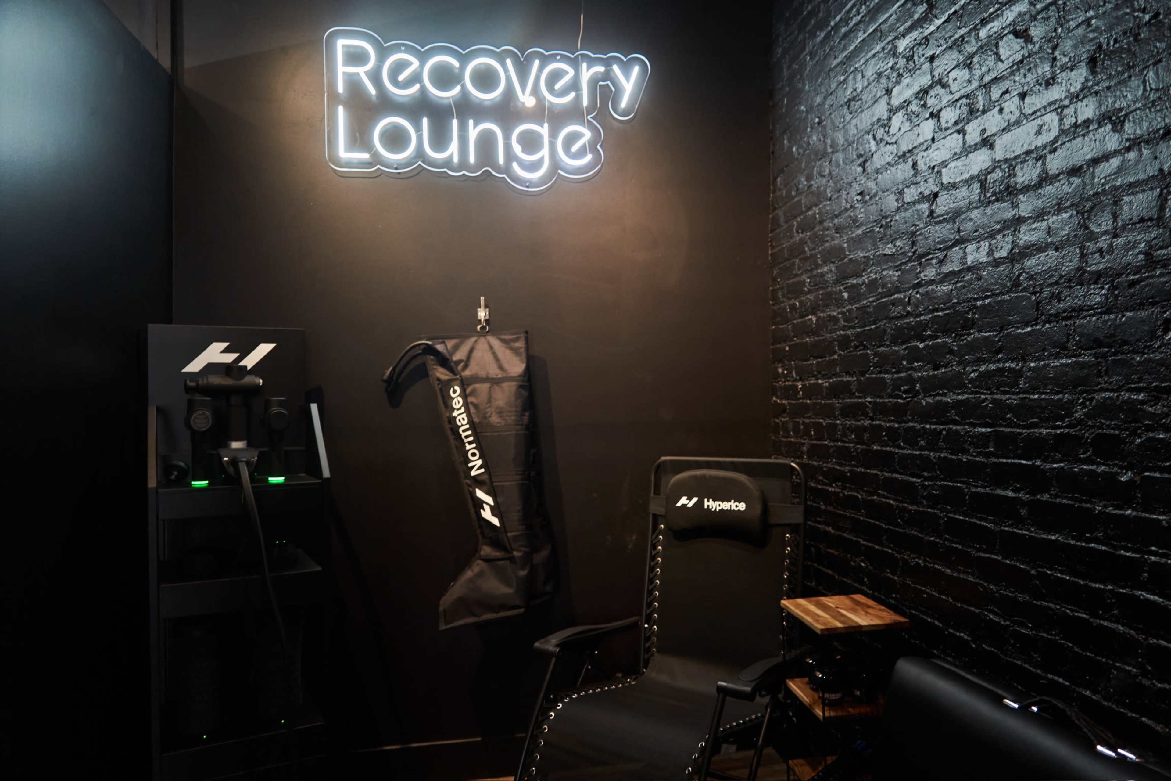 The image shows a small recovery lounge with a neon sign that reads "Recovery Lounge," a treatment chair, and equipment arranged against a black brick wall.