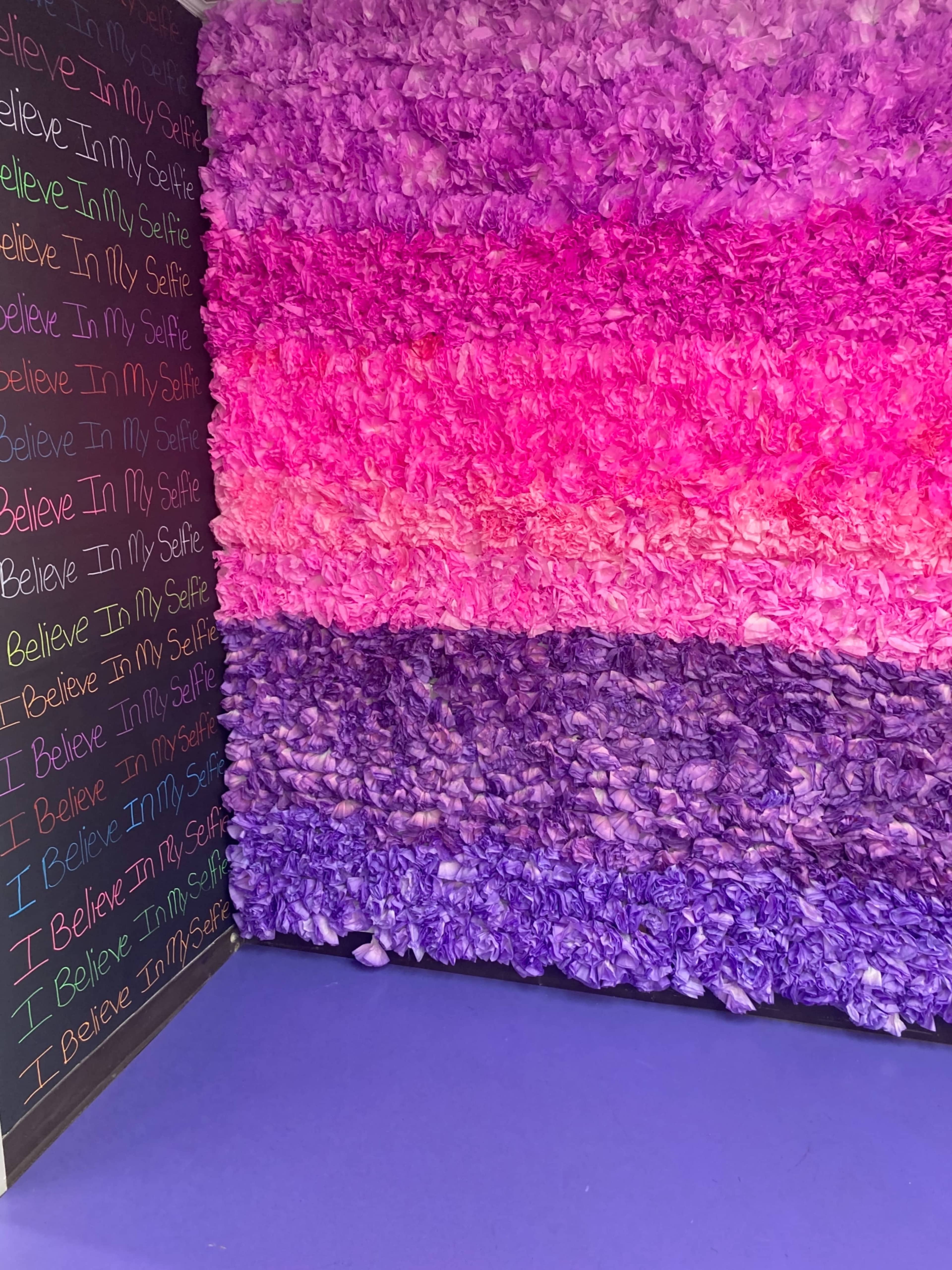 The image shows a wall composed of layered fabric flowers in shades of pink and purple, alongside a black wall featuring repetitive white text that says "I Believe In My Selfie."