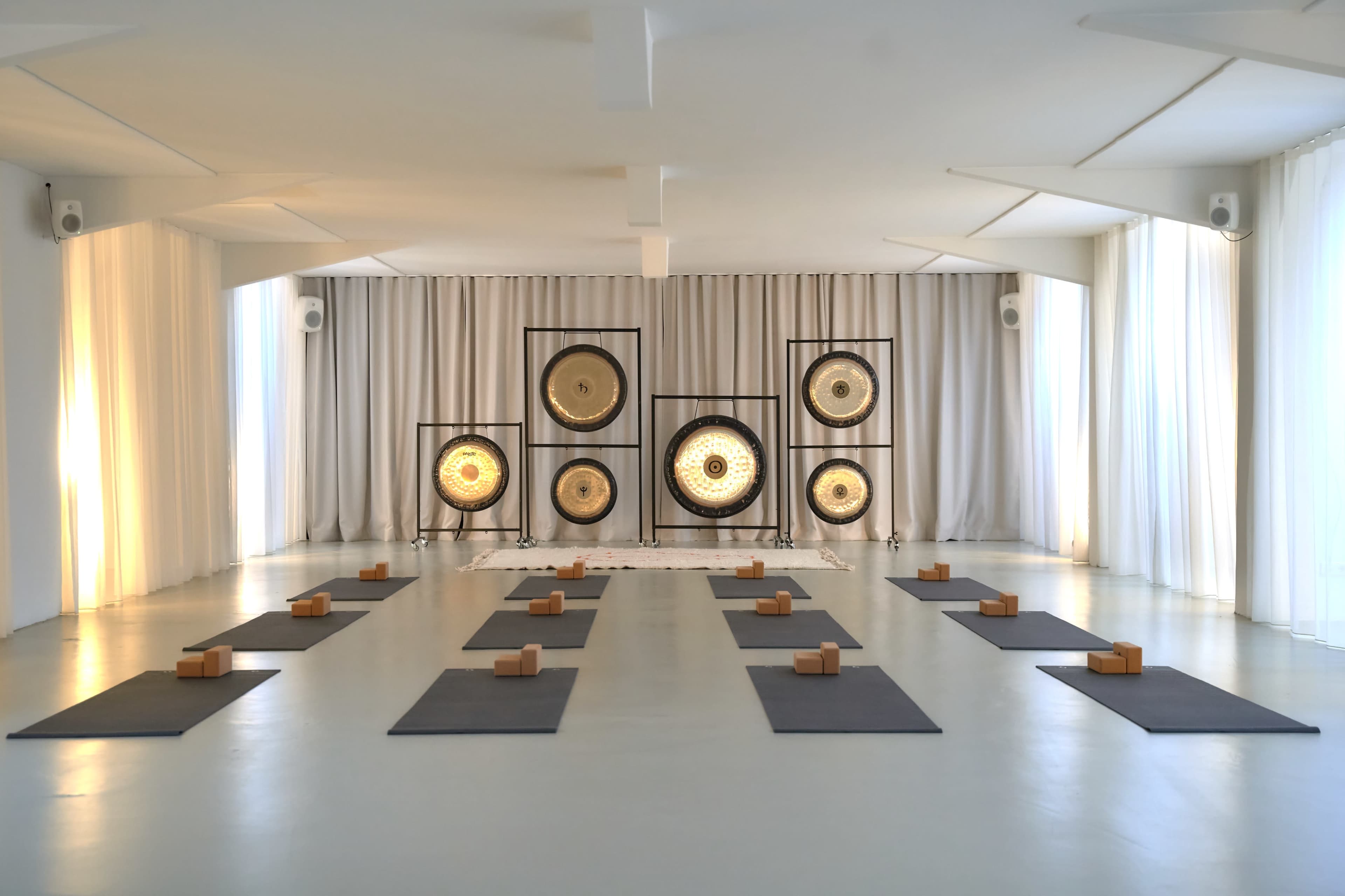 The image shows a spacious yoga studio with wooden blocks and mats arranged neatly on the floor, facing a wall adorned with large gongs.